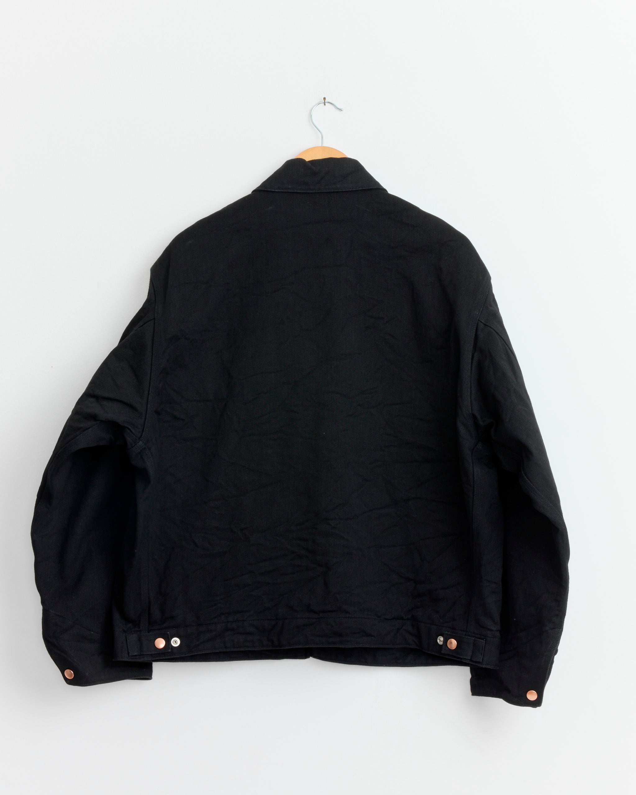 The Comoli Denim Zip Short Jacket in Black, photographed hanging on a wooden hanger against a white wall, features long sleeves and metal buttons at the hem. Made in Japan for exceptional craftsmanship.