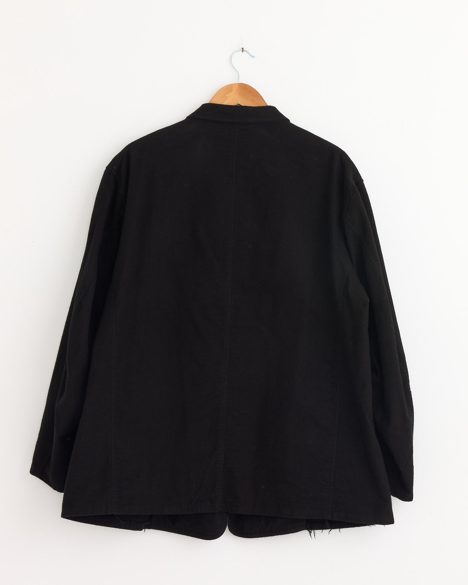 A Comoli Cotton Flannel Jacket in Black, made in Japan, hangs on a wooden hanger against a plain white wall, displayed from the back.