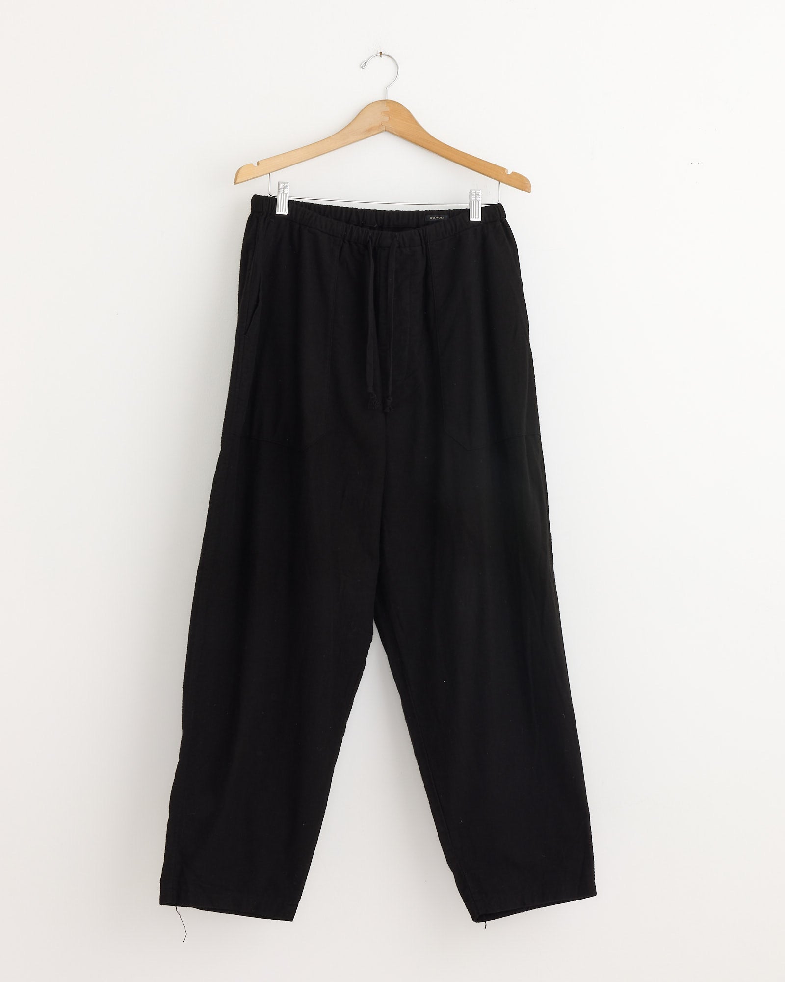 The Comoli Flannel Tapered Pant in Black, featuring an elastic waistband and crafted from soft cotton flannel in Japan, is displayed on a wooden hanger against a plain white wall.