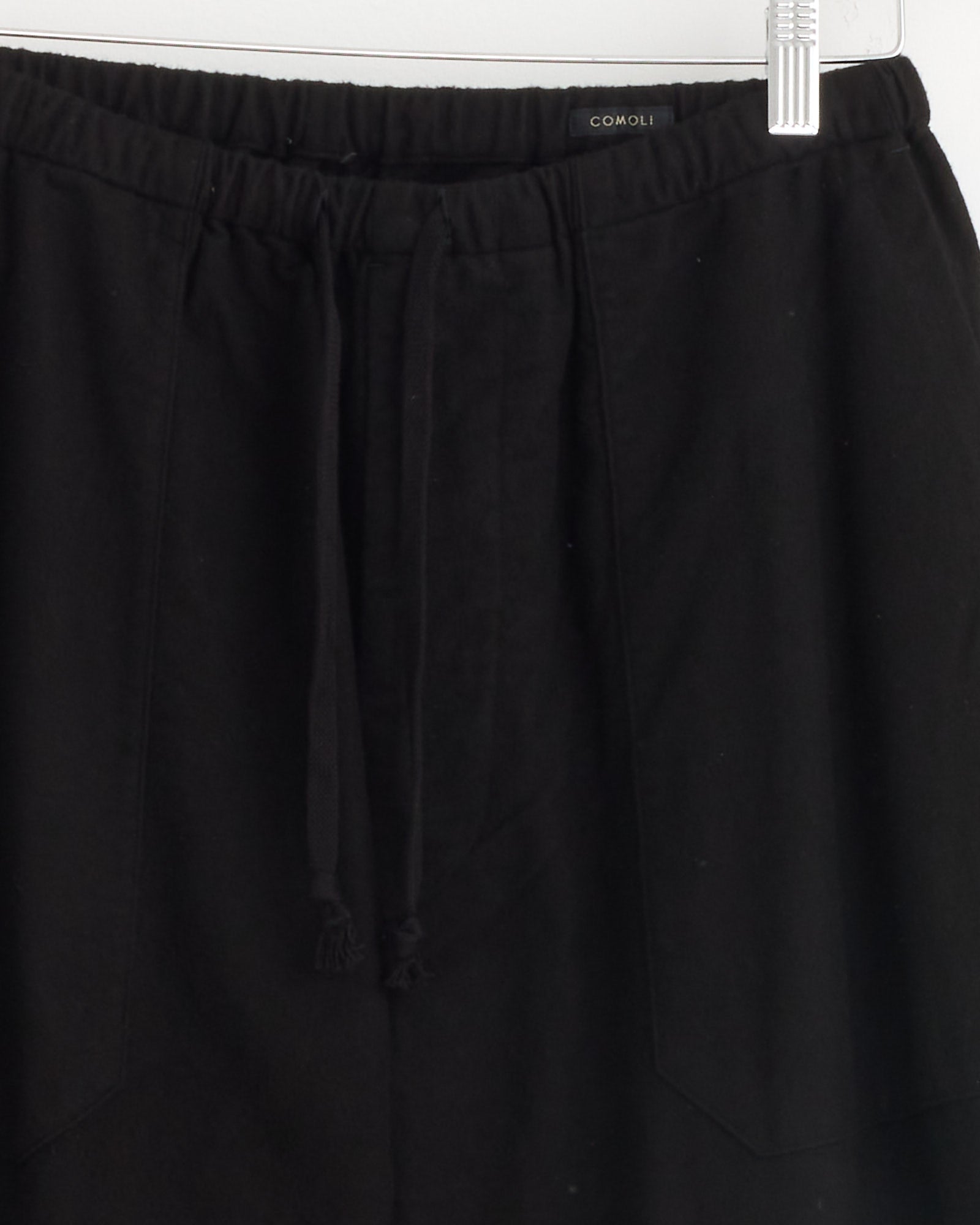 The Comoli Flannel Tapered Pant in Black, made in Japan, features an elastic waistband and visible brand label at the waist and is displayed on a clear plastic hanger.