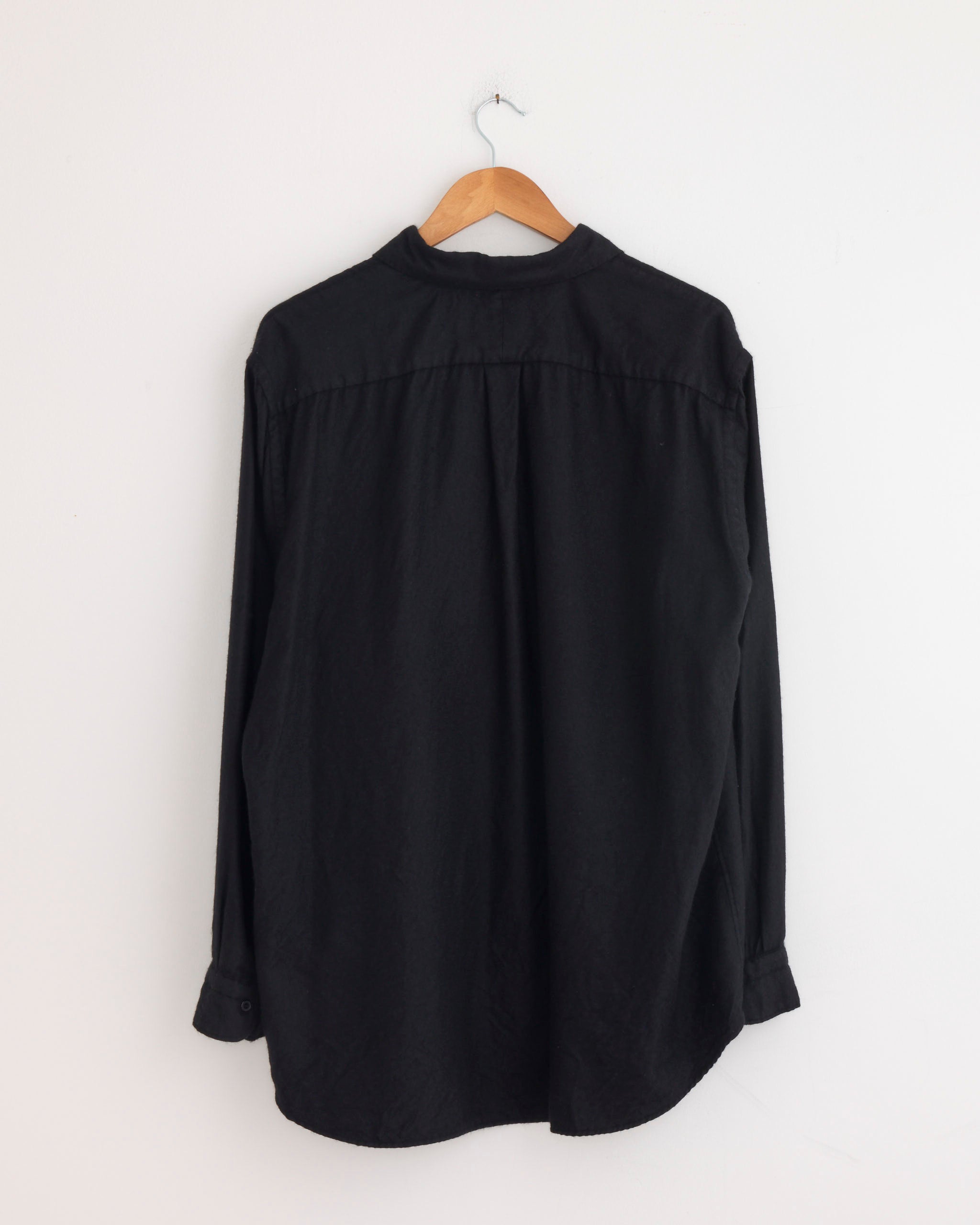 The Comoli Cashmere Silk Garment Dyed Shirt in Navy hangs on a wooden hanger against a plain white wall, displayed from the back.