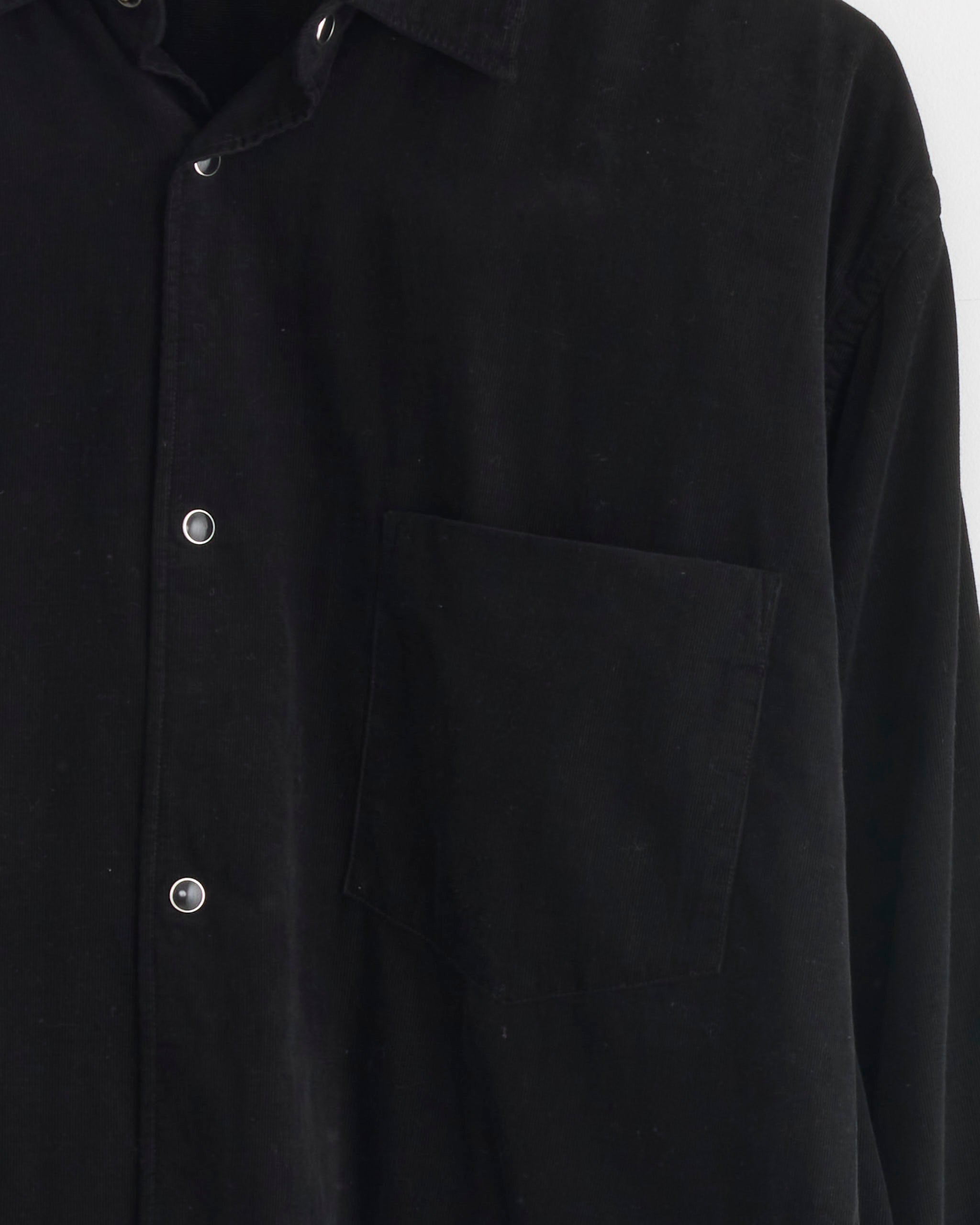 Close-up of the Corduroy Shirt in Black by Comoli, crafted from a soft cotton-cashmere blend with silver snap buttons and a single chest pocket. Only part of the collar and upper torso are shown.