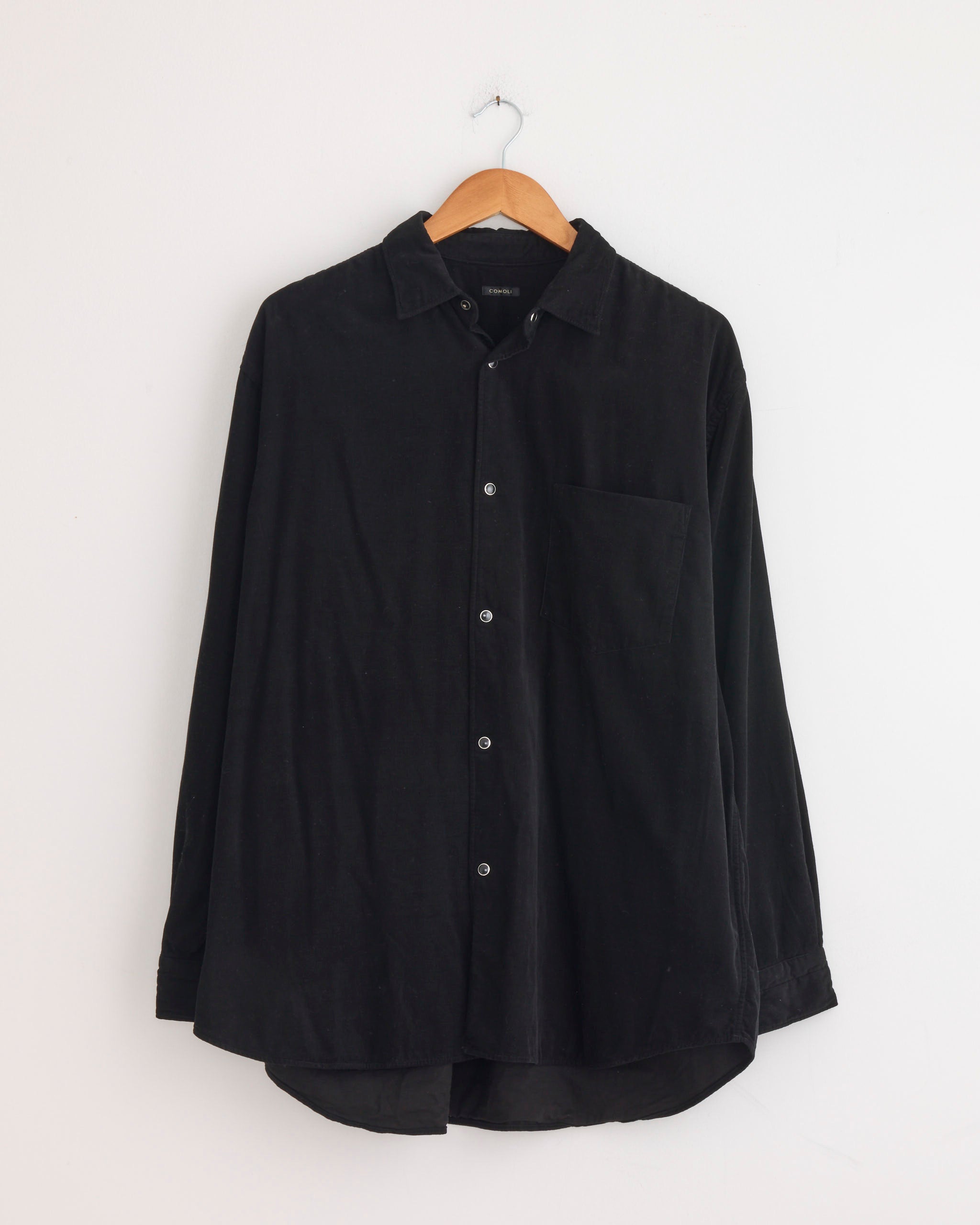 The Comoli Corduroy Shirt in Black, featuring long sleeves, a collar, and chest pocket, hangs on a wooden hanger against a plain white wall.