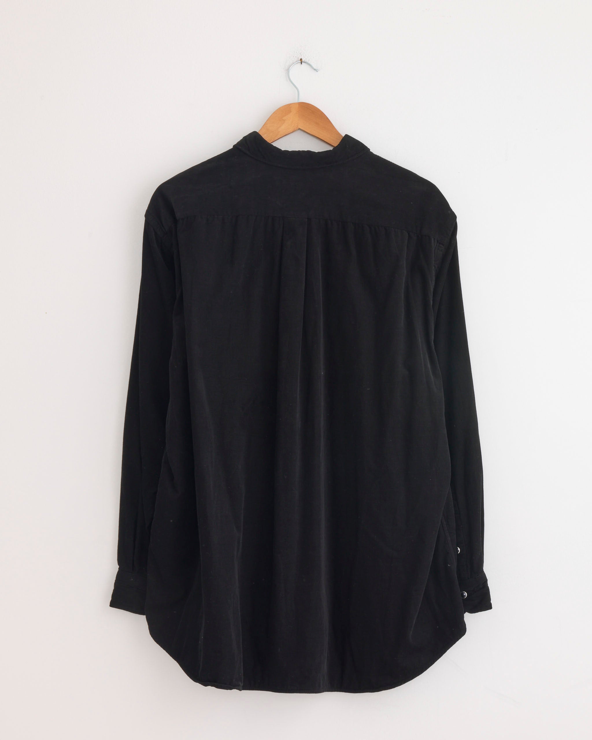 A Comoli Corduroy Shirt in Black hangs on a wooden hanger against a plain white wall, shown with the back facing forward.