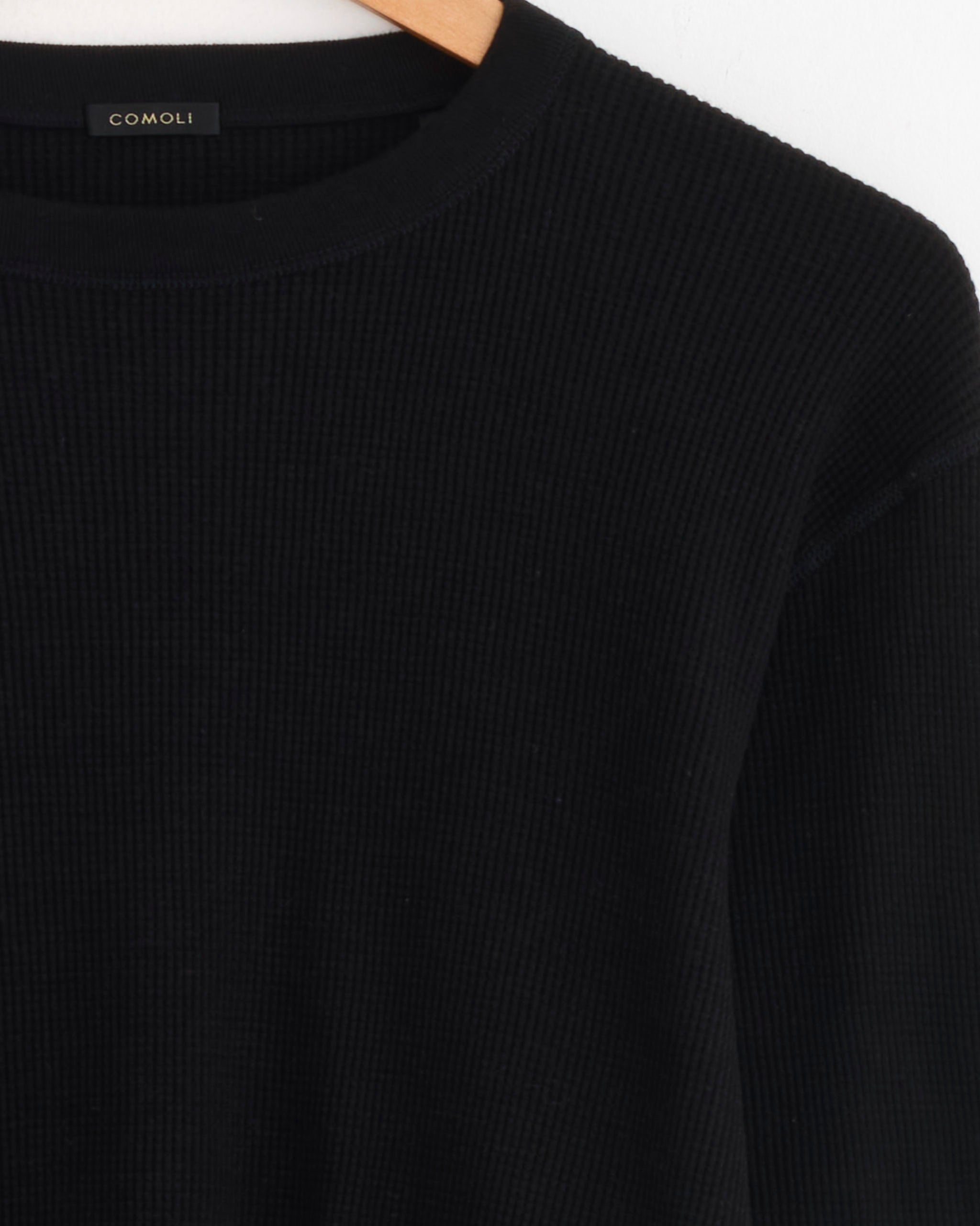 A close-up of the Comoli Cotton Cashmere Waffle L/S Crew in Black on a hanger, featuring the COMOLI label inside the collar.