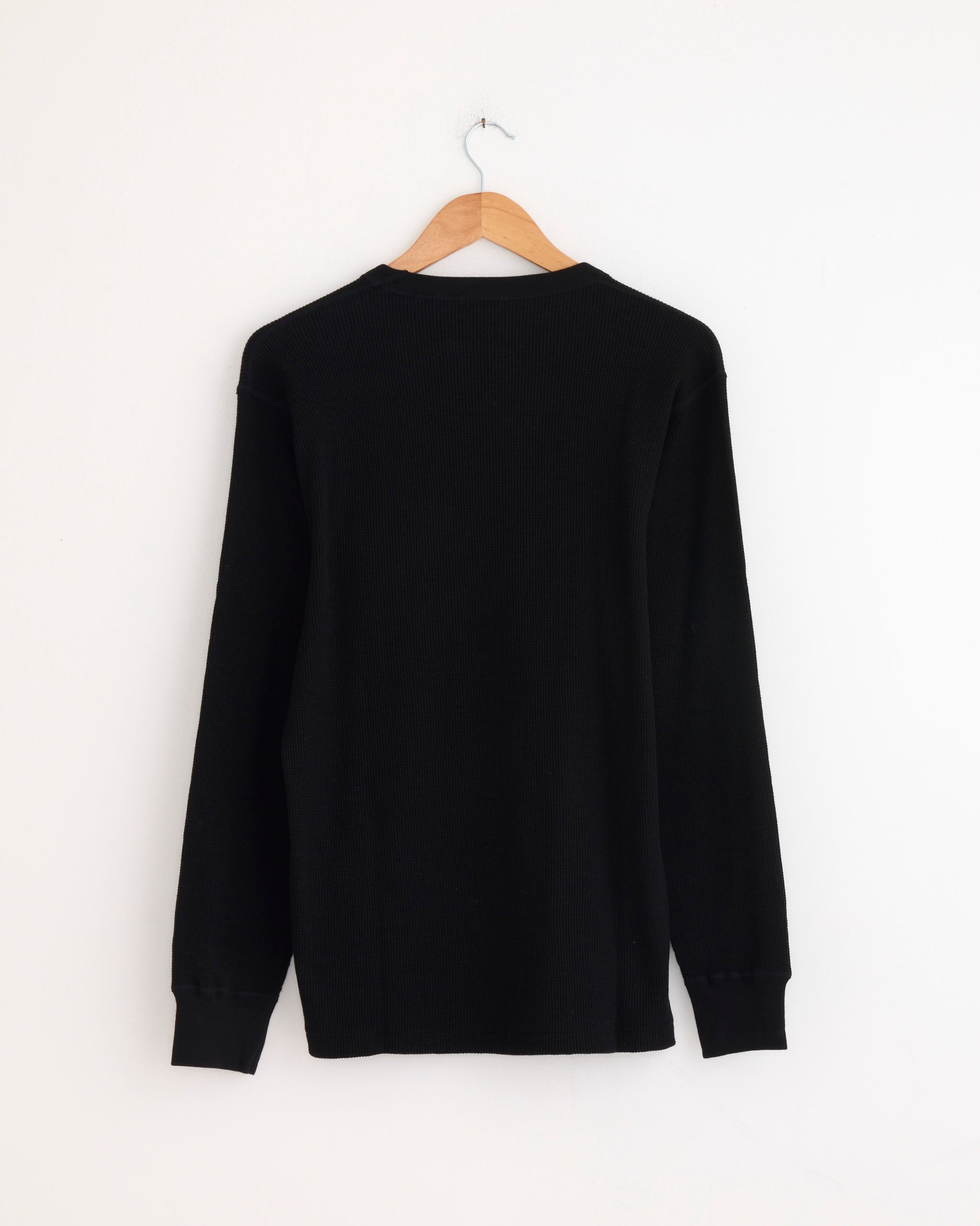 A Comoli Cotton Cashmere Waffle L/S Crew in Black hangs on a wooden hanger against a white wall, displayed from the back.