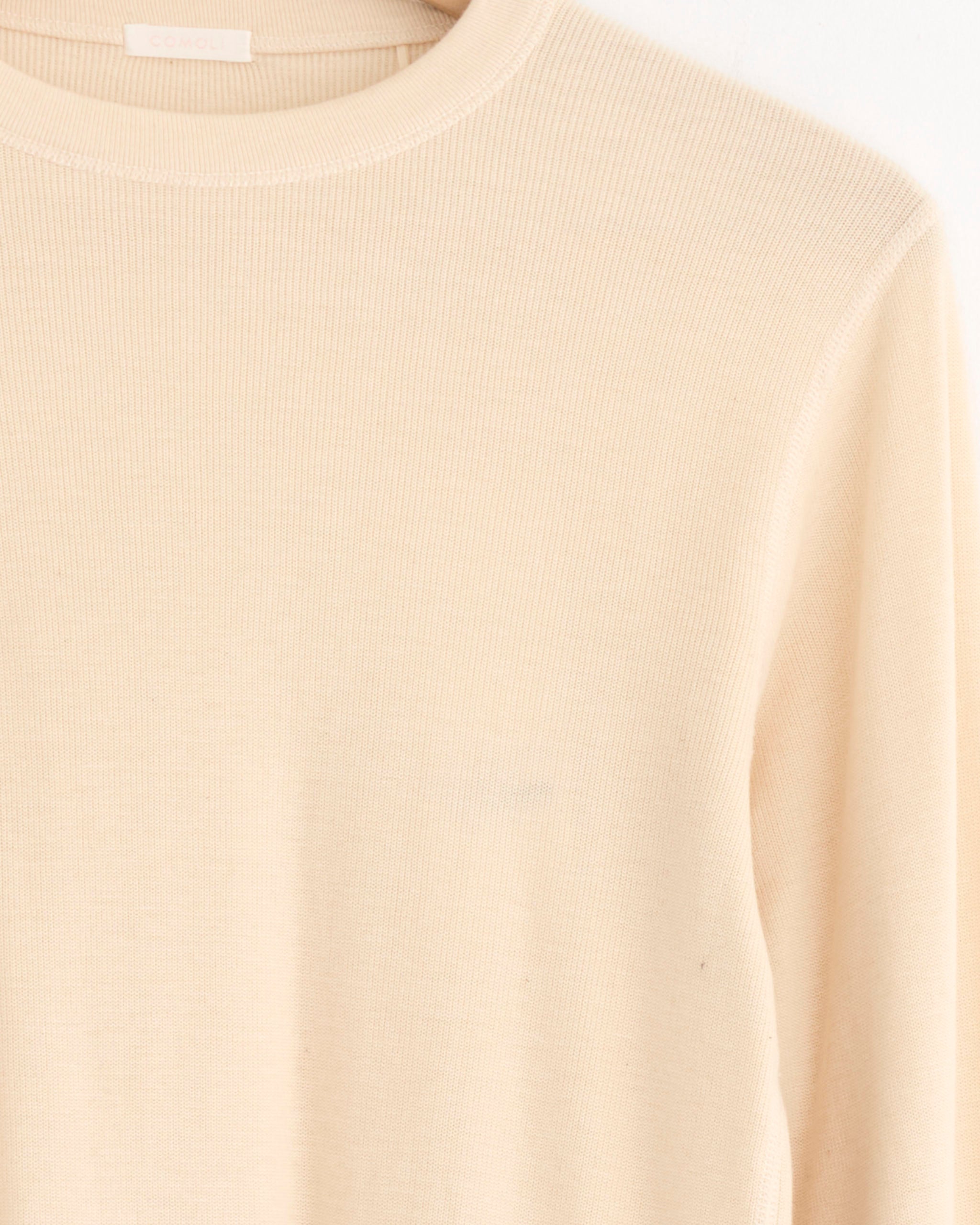 Close-up of the Comoli Cotton Cashmere Base Layer L/S Crew in Ecru, showcasing its ribbed-knit texture and crew neckline—an ideal lightweight layer against a white background.
