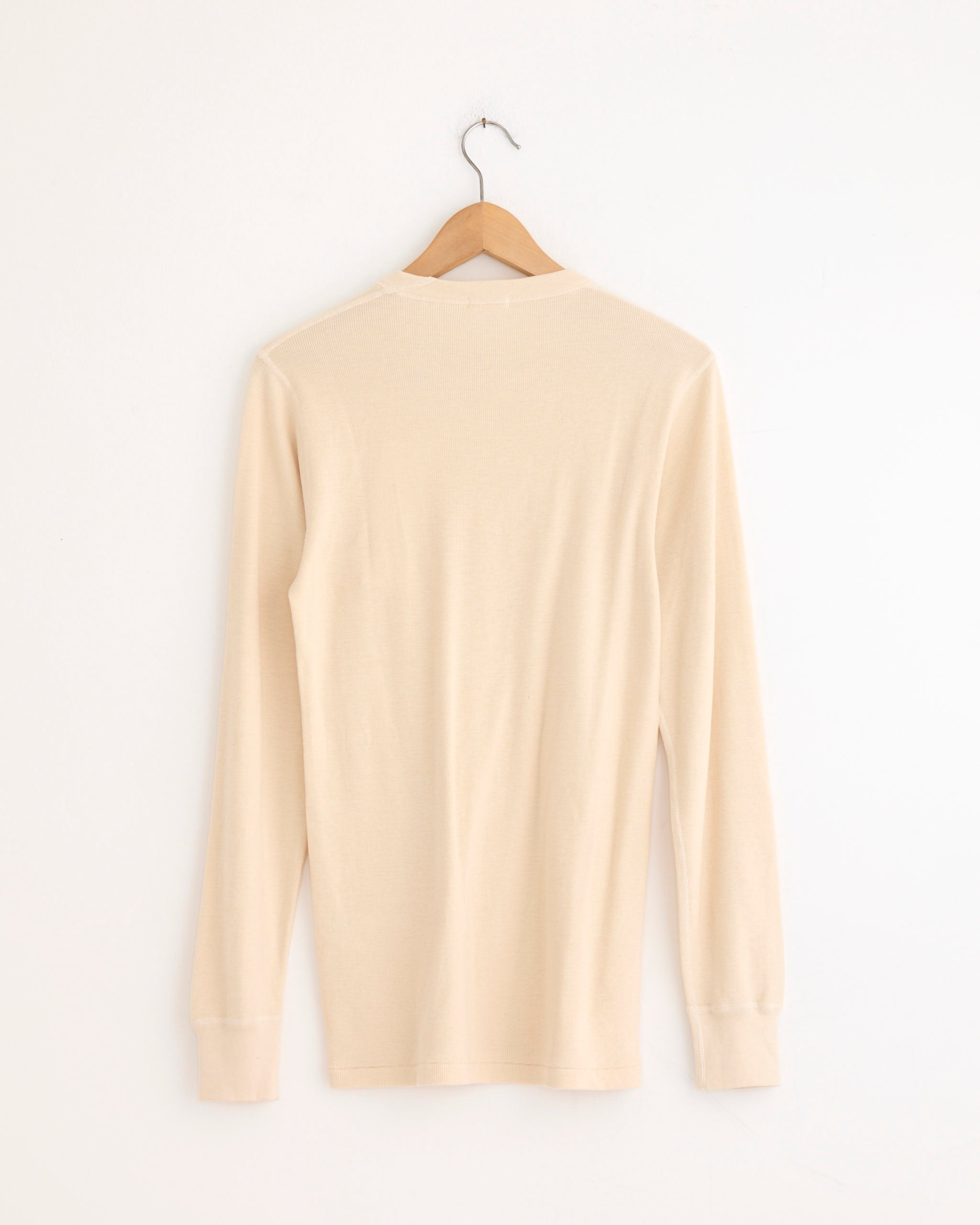 A Comoli Cotton Cashmere Base Layer L/S Crew in Ecru hangs on a wooden hanger against a plain white wall, displayed from the back.