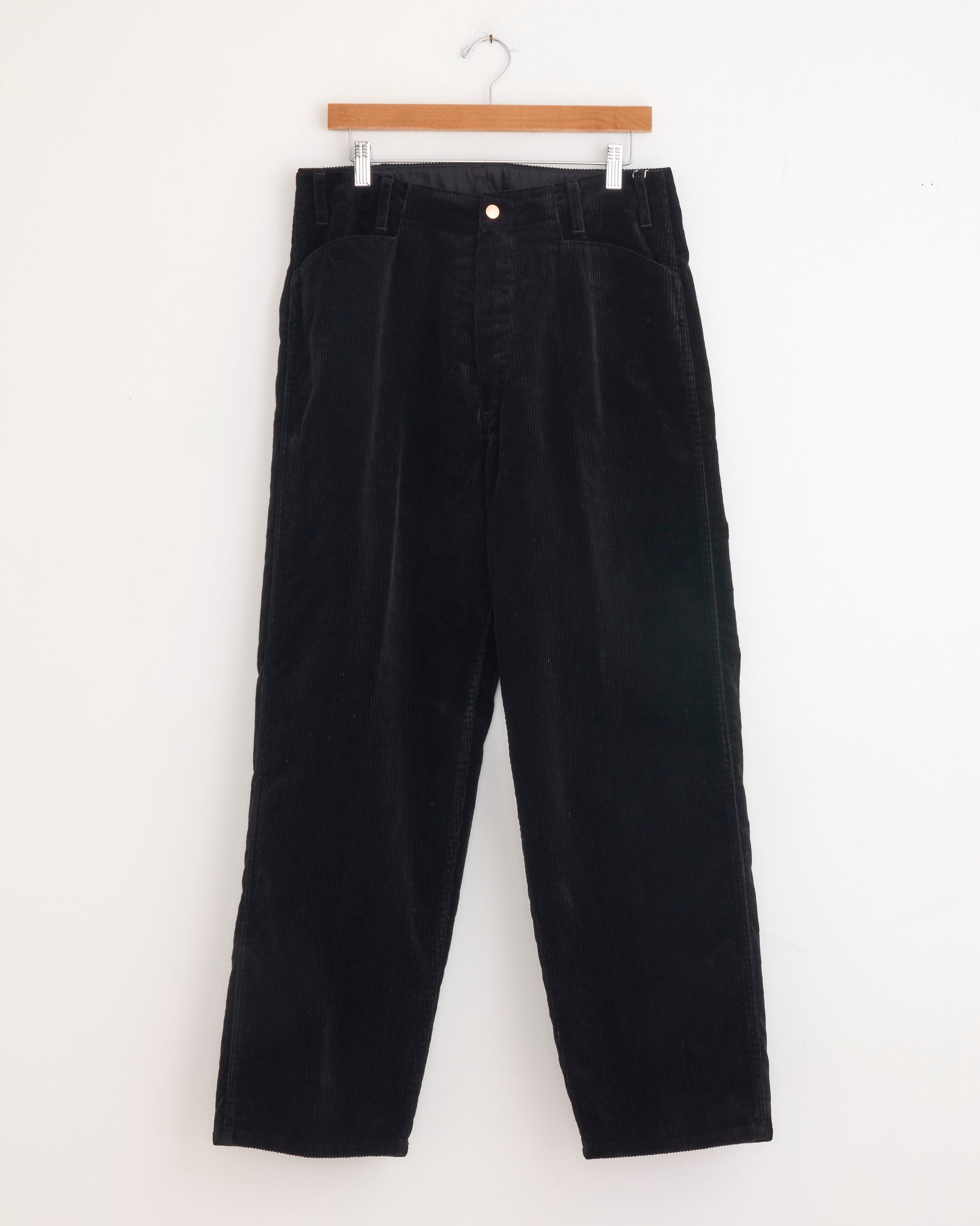 A pair of Comoli Corduroy Work Pant in Black, crafted from 100% cotton corduroy, hangs on a wooden hanger against a plain white background.