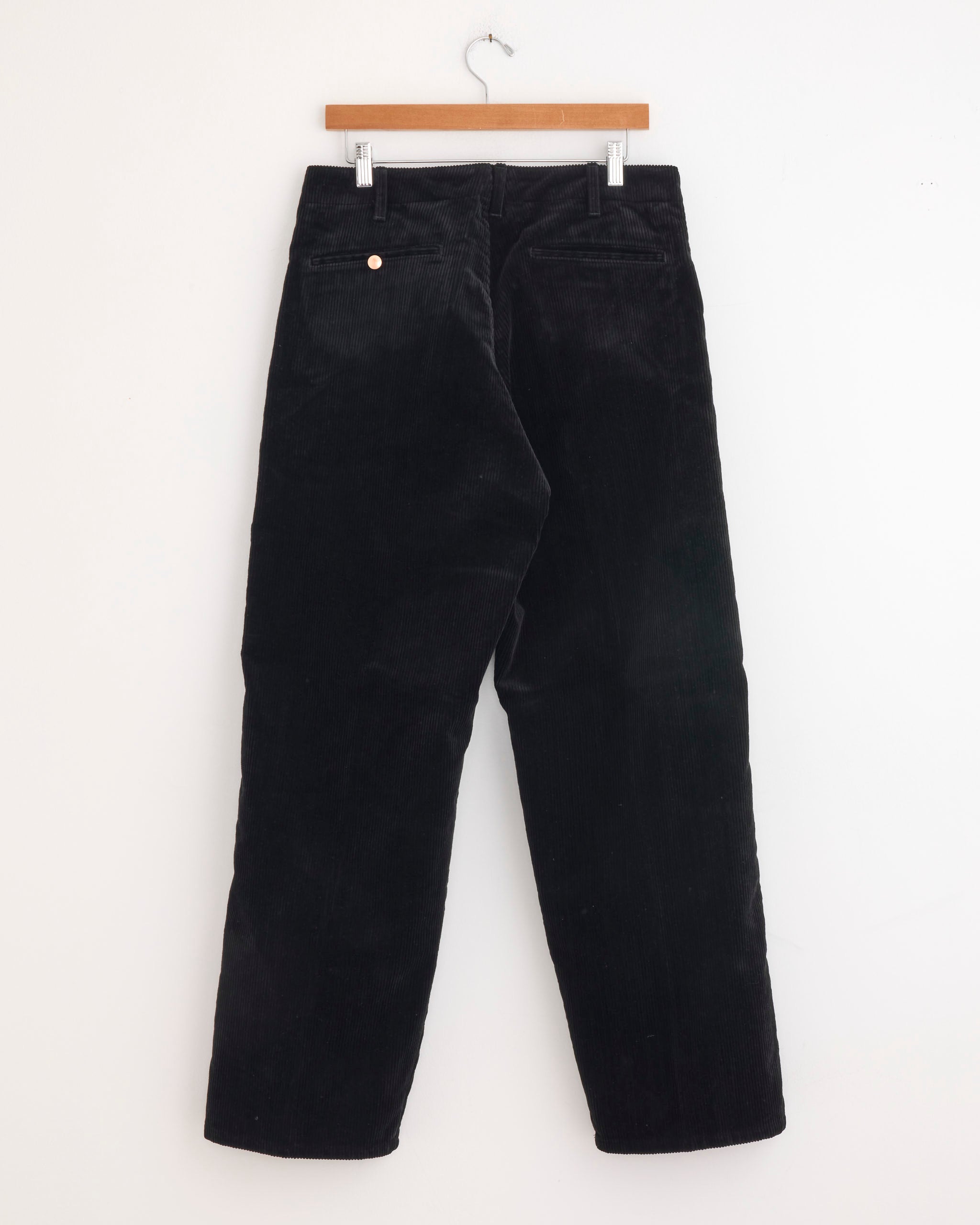 The Comoli Corduroy Work Pant in Black, made from 100% cotton with a straight-leg cut and single back pocket, hangs on a wooden hanger against a plain white background.