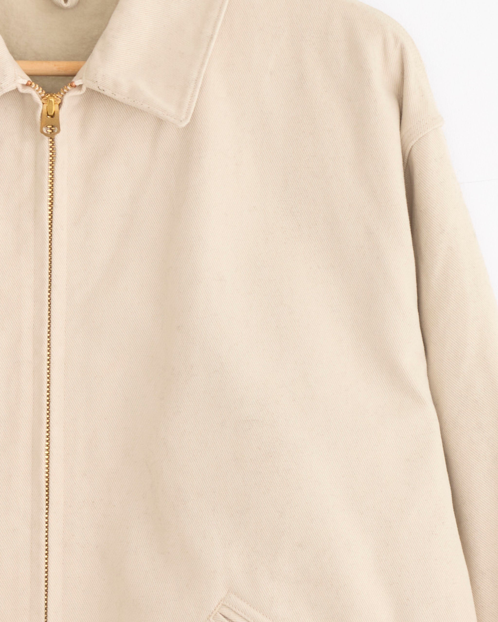A close-up of the Comoli Cotton Twill Zip Short Jacket in Ivory hanging on a wooden hanger, highlighting its collar, gold zipper, and front pocket against a plain white background.