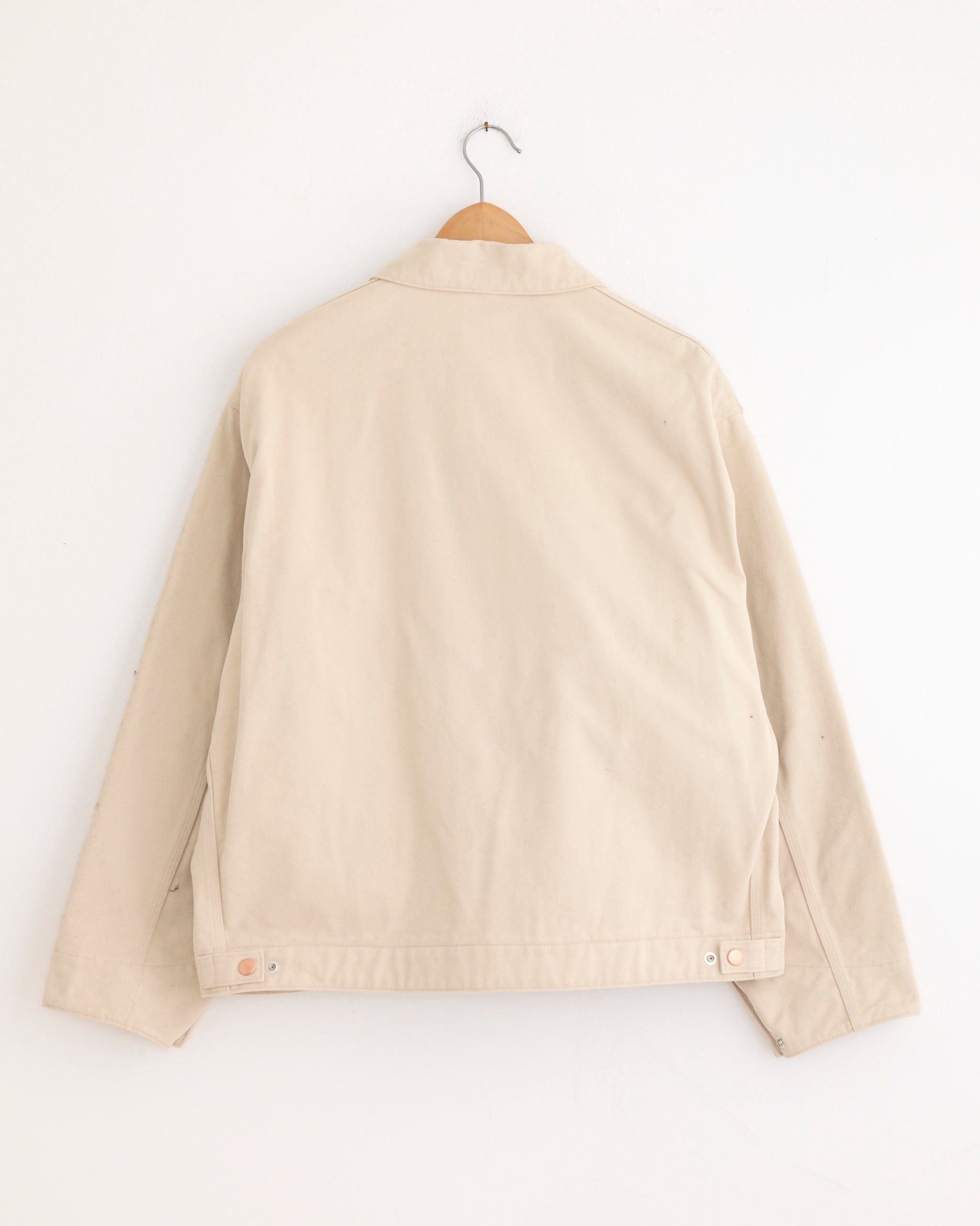 The Comoli Cotton Twill Zip Short Jacket in Ivory hangs on a wooden hanger against a white wall, displayed from the back. This ivory cotton jacket features long sleeves and adjustable button tabs at the hem.