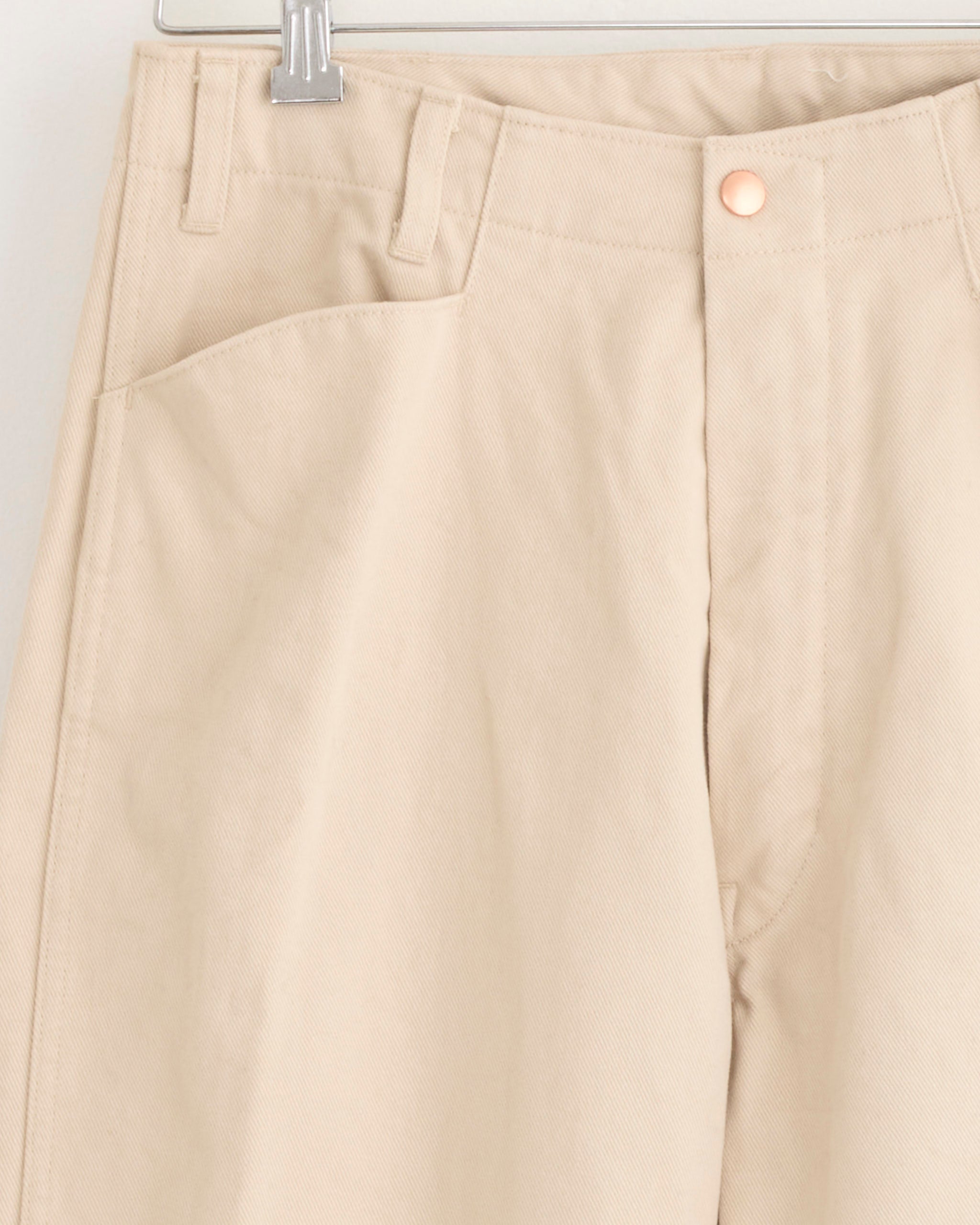 Close-up of the Comoli Cotton Twill Work Pant in Ivory hanging on a hanger, highlighting the waistband, belt loops, front pocket, and button closure. Crafted from heavyweight cotton twill with a sturdy texture and wide straight silhouette.