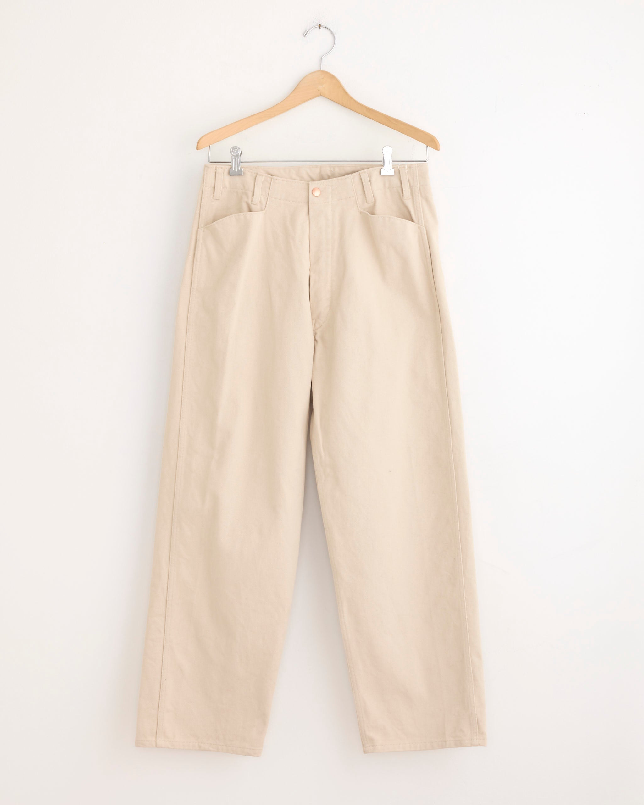 The Comoli Cotton Twill Work Pant in Ivory hangs on a wooden hanger against a white wall. Made from heavyweight cotton twill, these high-waisted pants feature a wide straight silhouette, front pockets, and belt loops.
