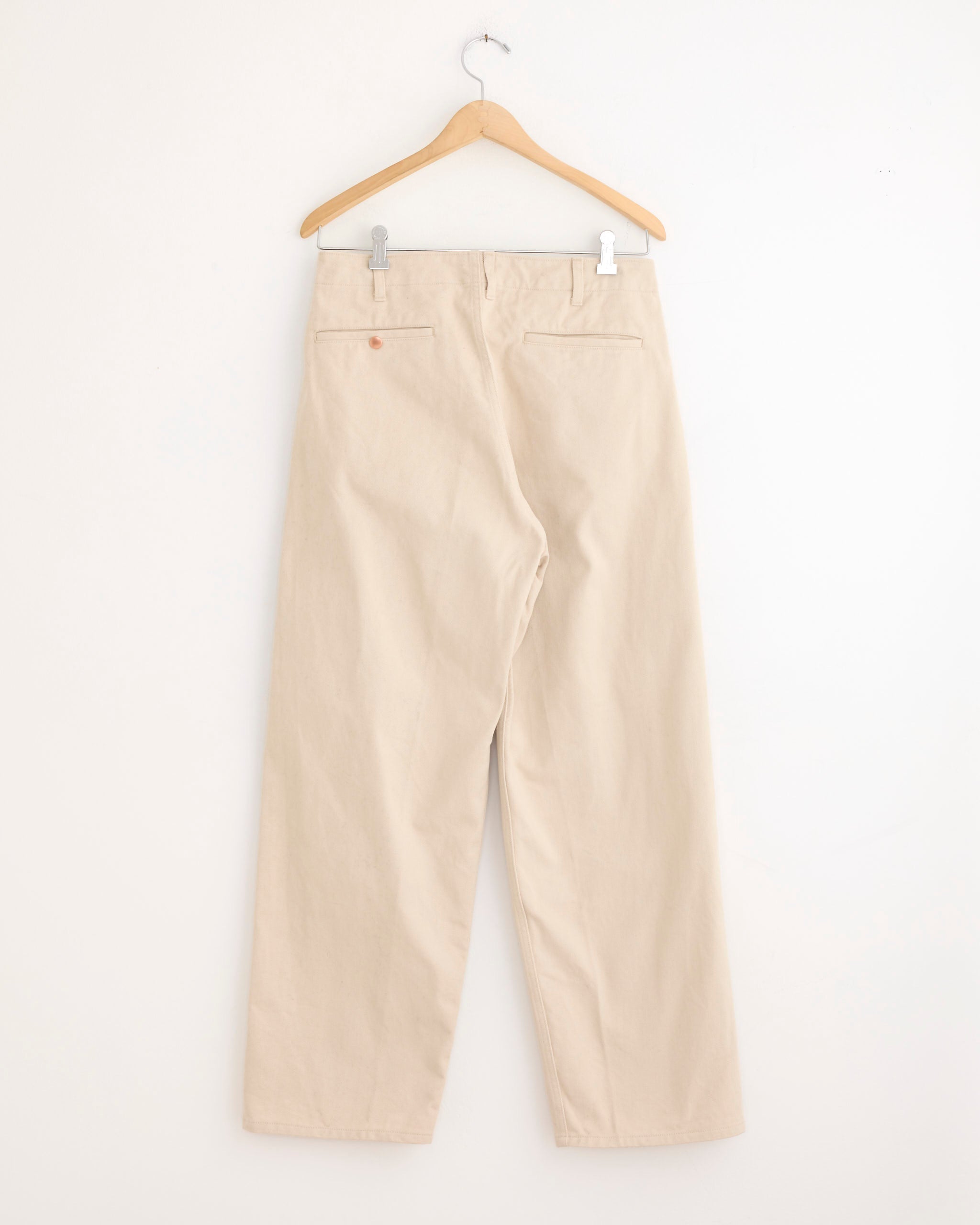 A pair of Comoli Cotton Twill Work Pant in Ivory, made from heavyweight cotton twill with a wide straight fit, hang on a wooden hanger before a plain white wall. The back has two pockets, including one with a button closure.