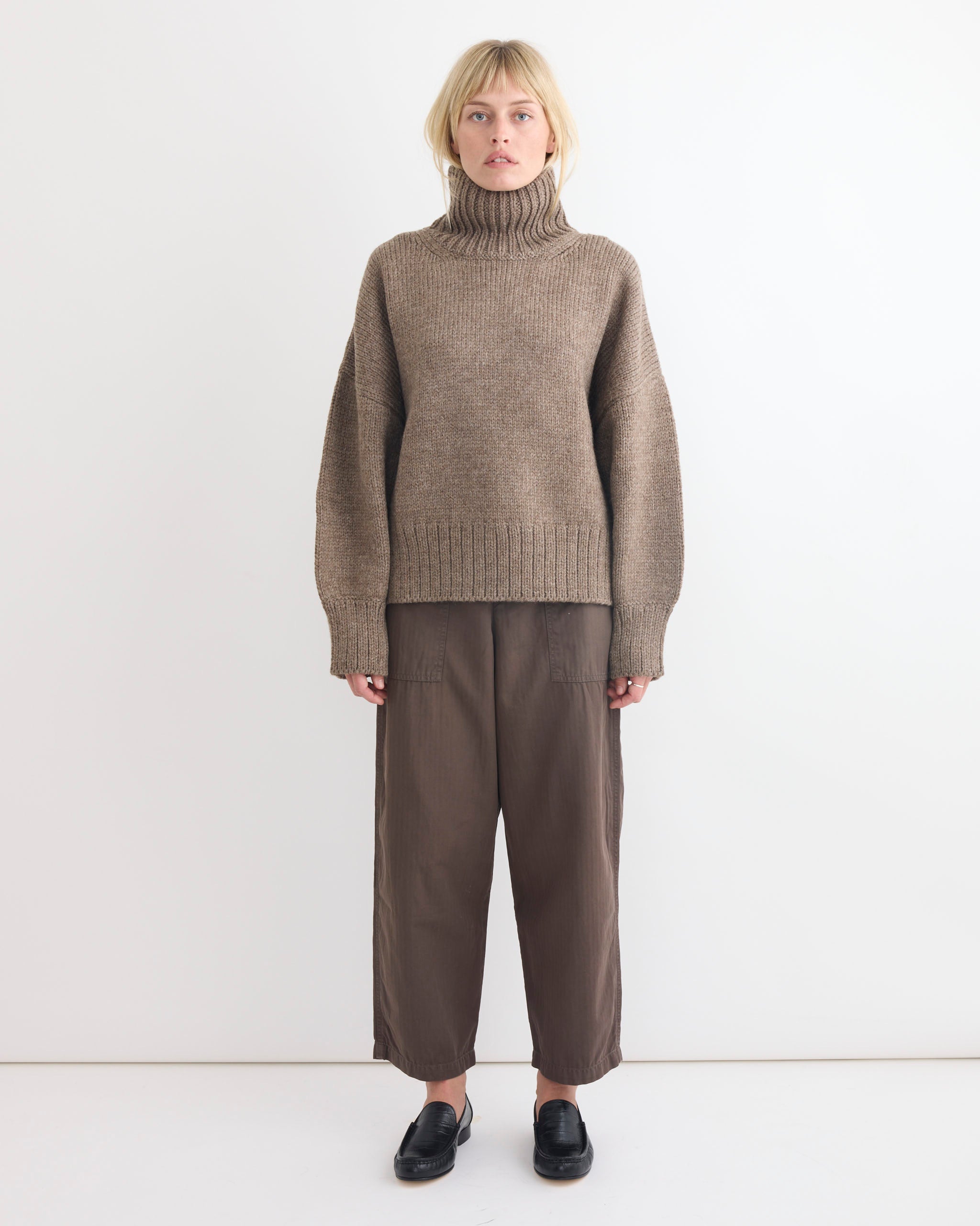 A person stands against a plain white wall wearing the oversized Shaina Mote Rhys Sweater in Tundra, loose brown pants, and black slip-on shoes. Their expression is neutral, hands resting by their sides.
