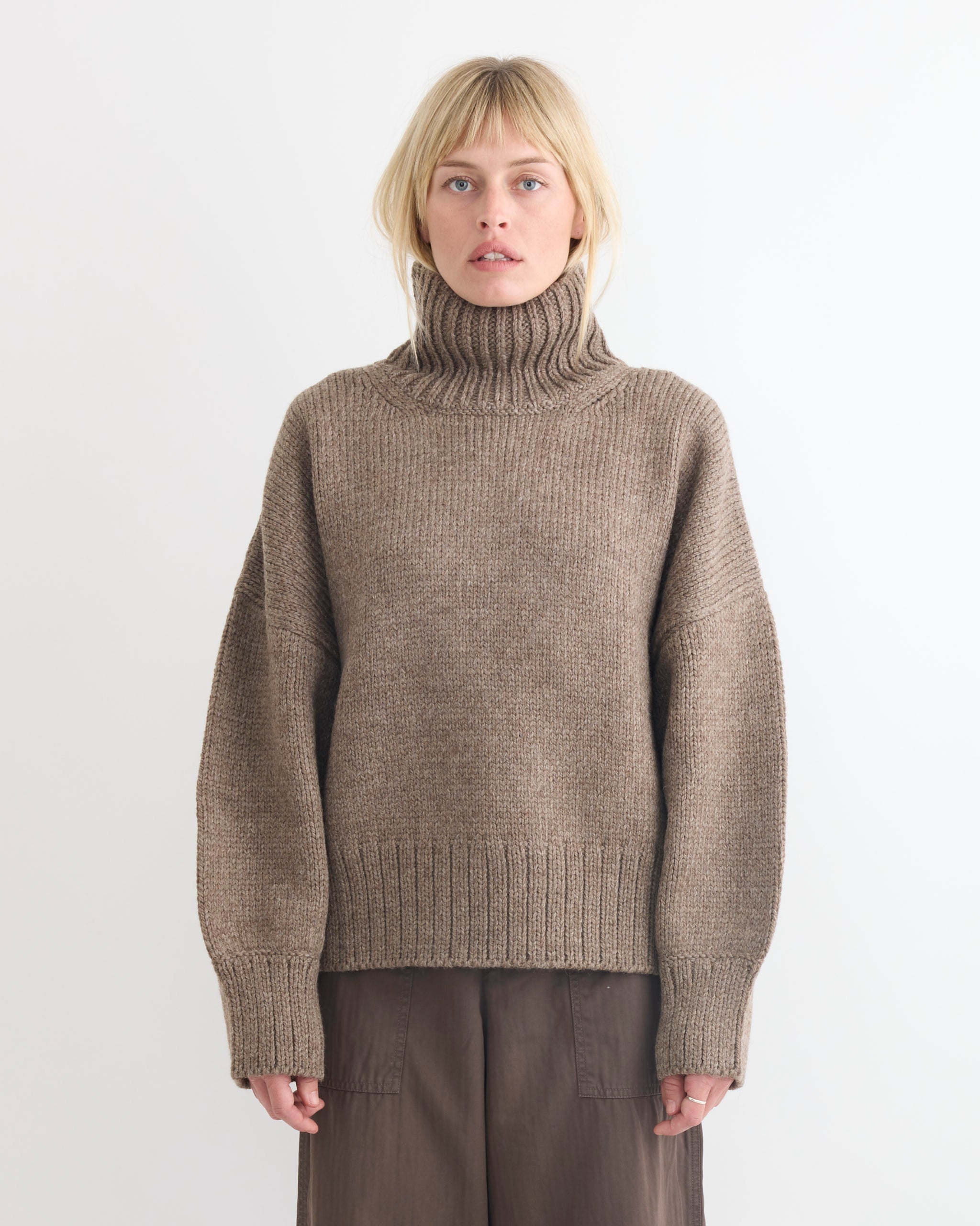 A woman with blonde hair wears the Shaina Mote Rhys Sweater in Tundra, paired with loose brown pants. She stands against a white background, looking directly at the camera with a neutral expression.