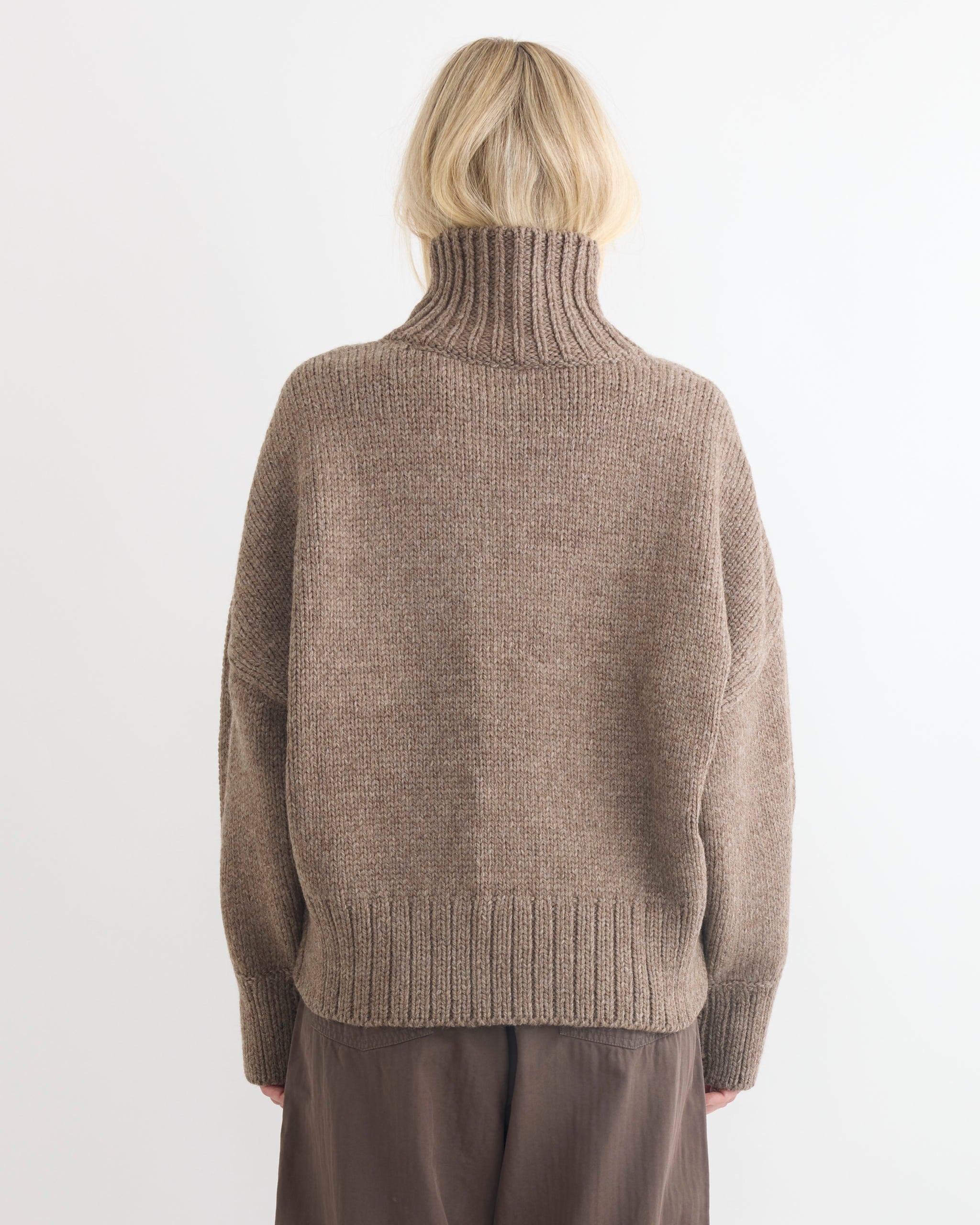 A person with blonde hair, seen from behind, wears the Rhys Sweater in Tundra by Shaina Mote and loose dark brown pants, standing against a plain white background.