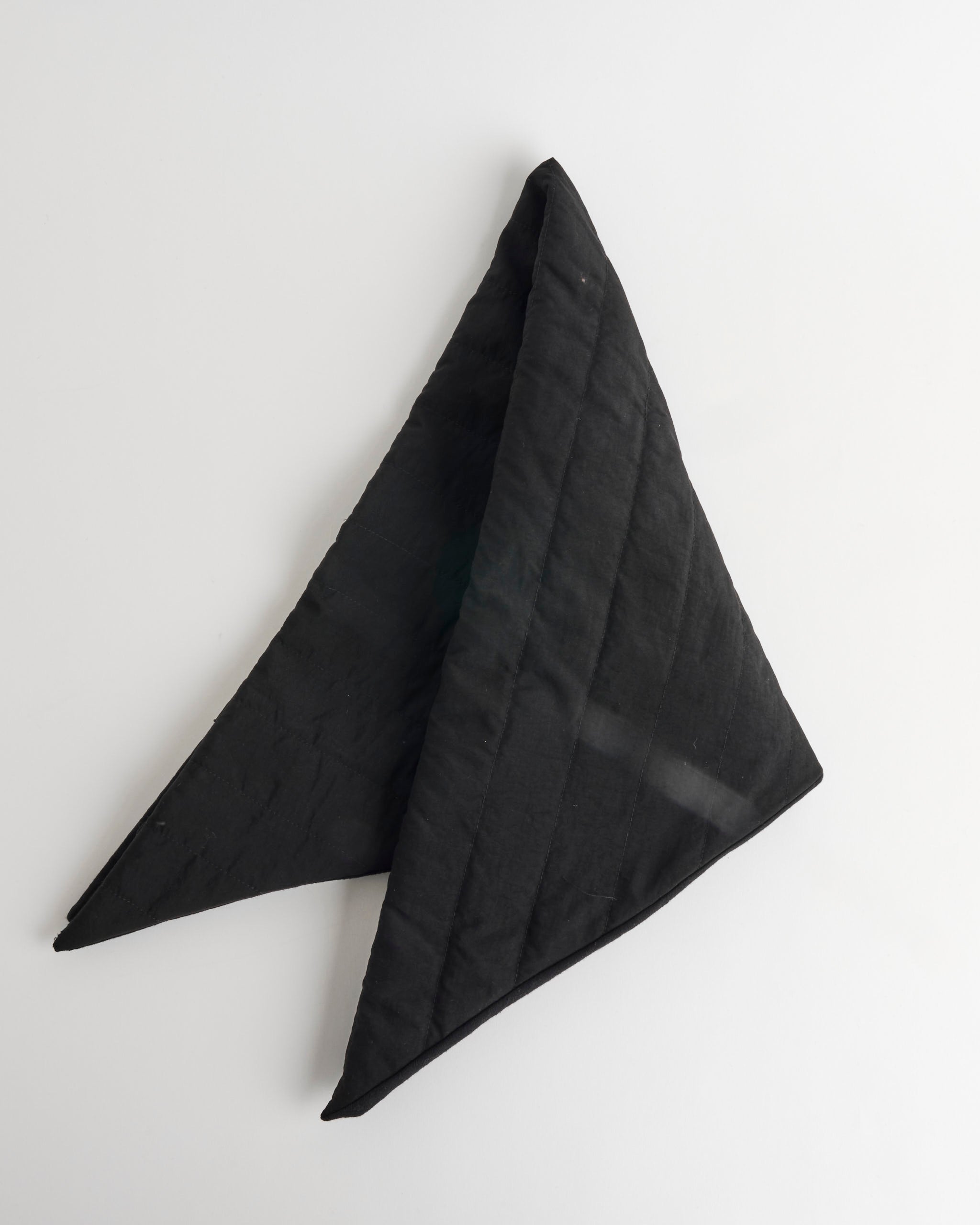 The Clyde Puff Scarf, a black, triangular quilted piece, is displayed flat on a white surface with two points extending left and right and the third pointing upward.