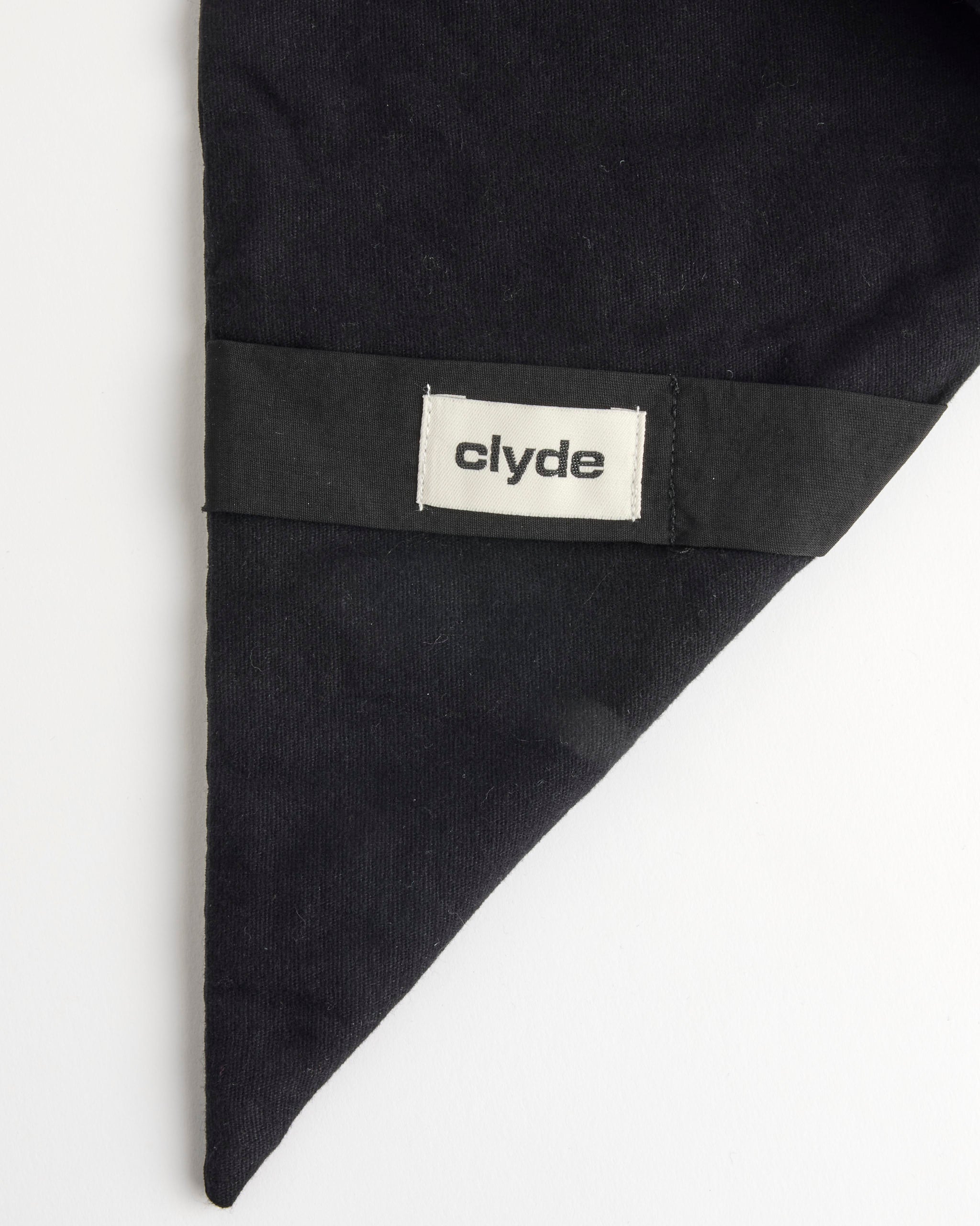 A black cotton flannel Puff Scarf by Clyde is shown on a white background. A rectangular white label with "clyde" in lowercase black letters is stitched onto a black band across the reversible neck scarf’s pointed end.