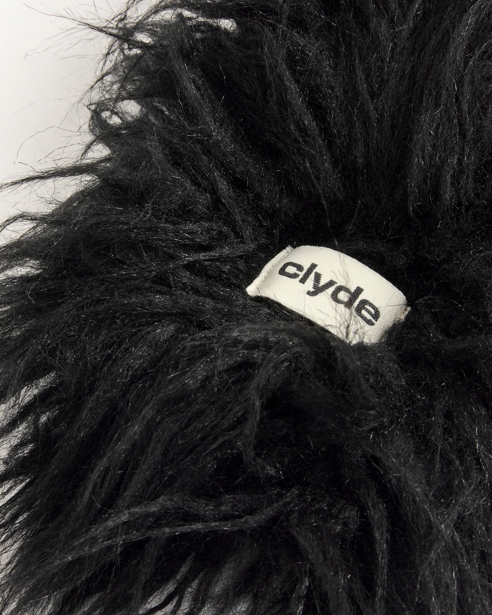 A close-up of the Sue Scarf by Clyde, a fluffy black faux fur scarf featuring a white fabric tag with "clyde" in lowercase black letters, set against a plain light background.