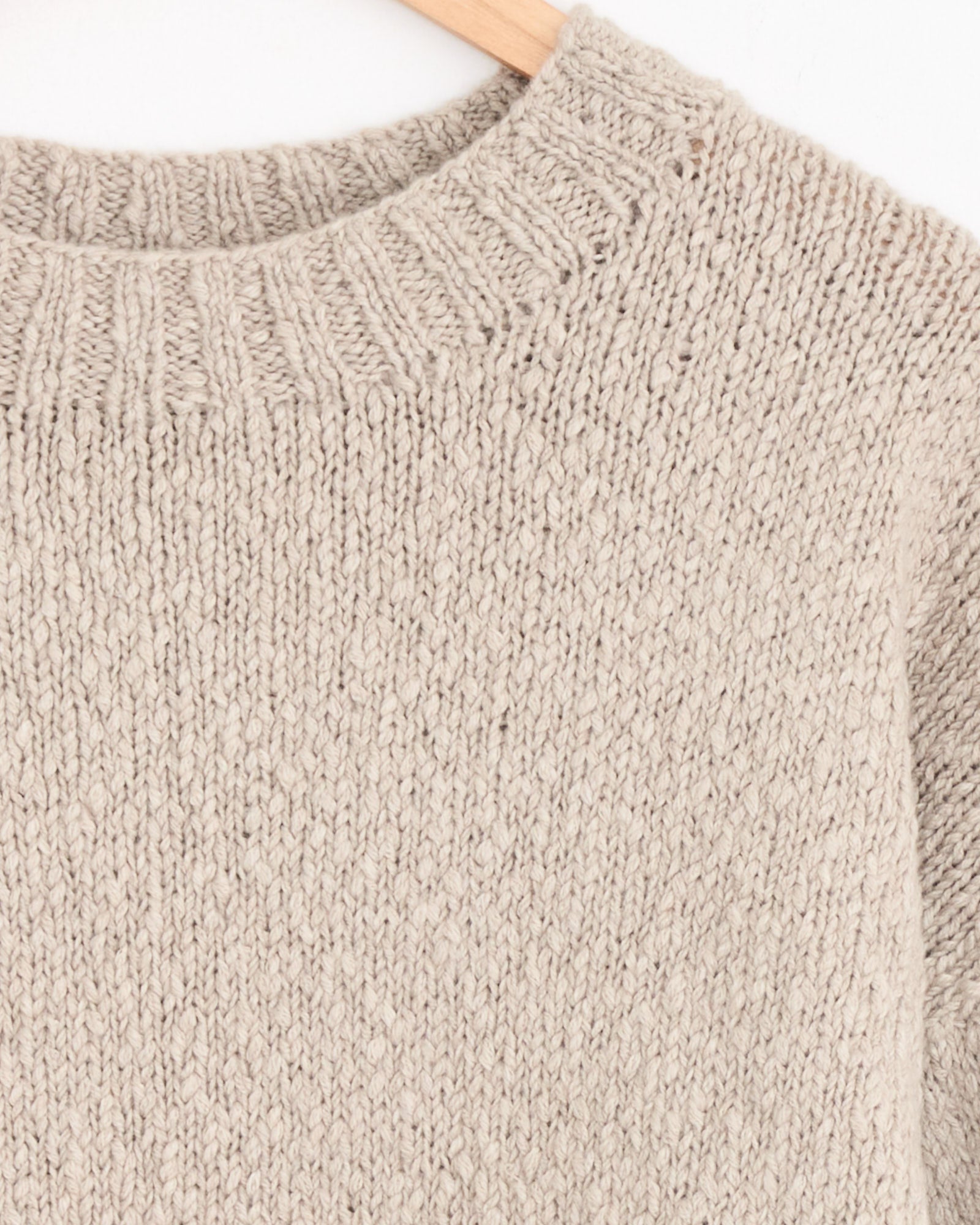 Close-up of the Cale Cashmere Hand Knit Sweater in Beige, featuring a thick ribbed neckline. Displayed on a wooden hanger against a white background, this sweater blends comfort and elegance.