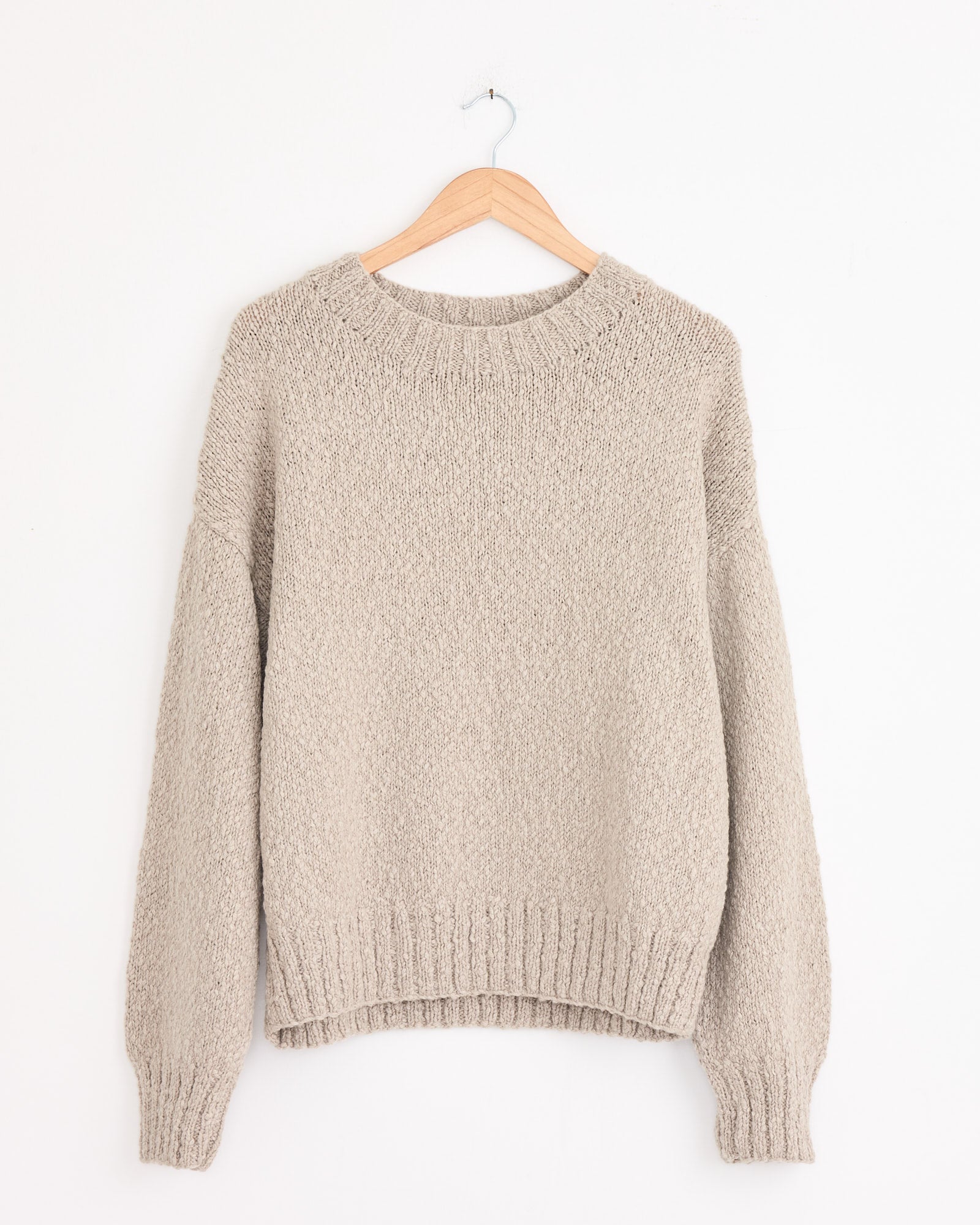The Cale Cashmere Hand Knit Sweater in Beige features long sleeves and ribbed collar, cuffs, and hem. Made in Japan, it's shown on a wooden hanger against a white wall.