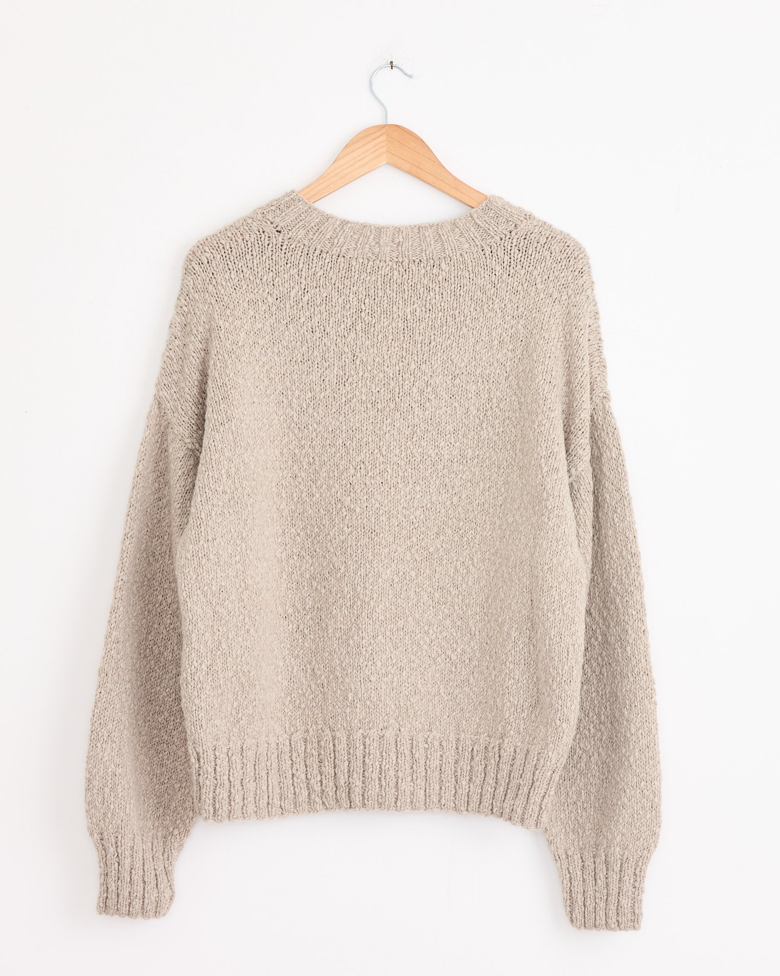 The Cale Cashmere Hand Knit Sweater in Beige, made in Japan, hangs on a wooden hanger against a white wall. It features long sleeves and ribbed hem, cuffs, and neckline for refined craftsmanship.