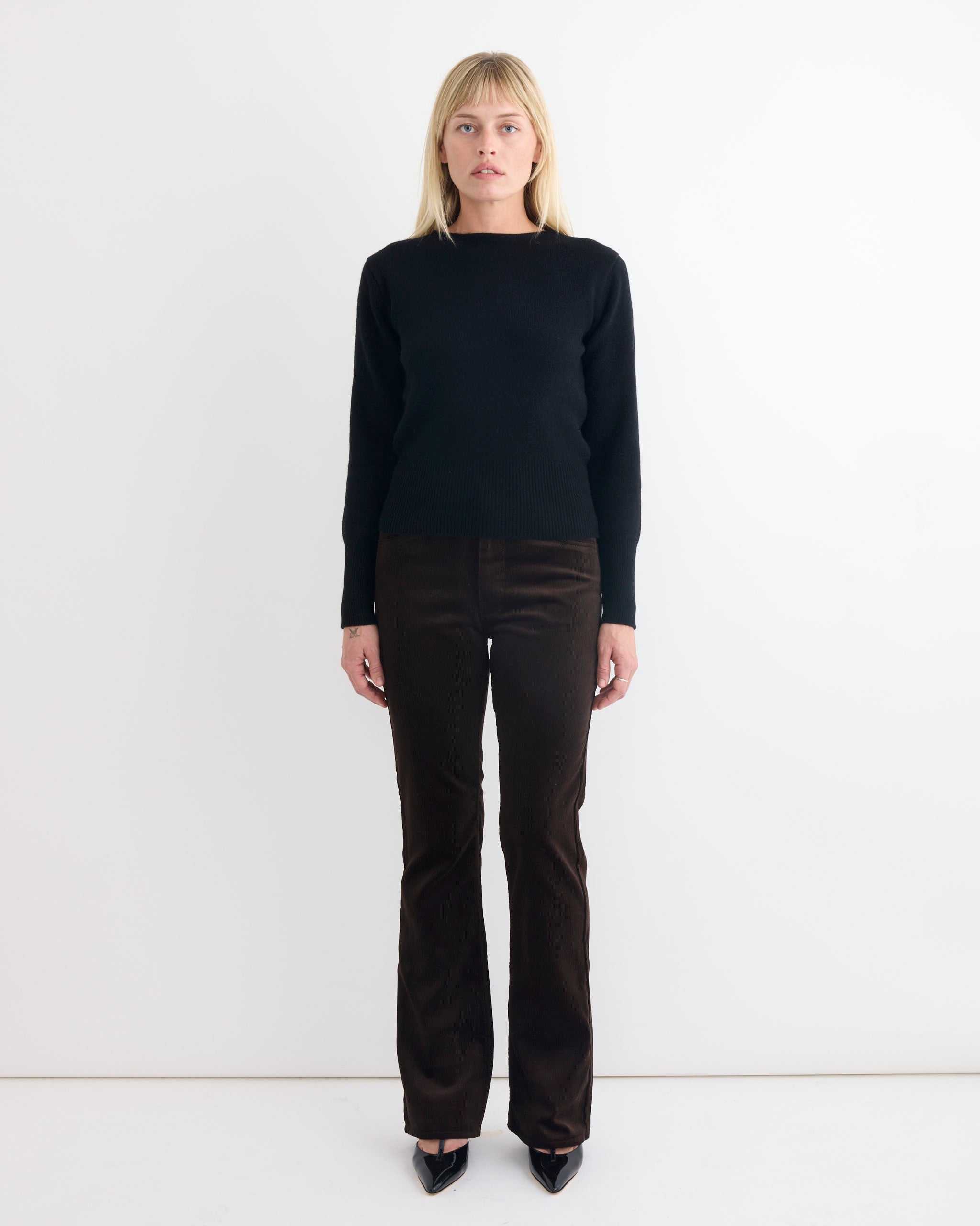 A woman with straight blonde hair stands facing forward against a white background, wearing a black long-sleeve sweater, Comoli Corduroy Pant in Brown made in Japan, and black pointed shoes.