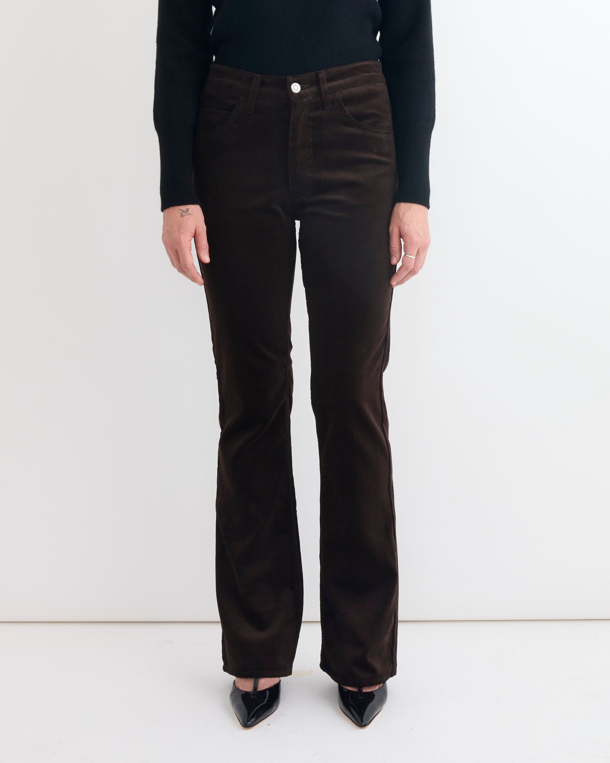 A person is shown from the shoulders down wearing Comoli's Corduroy Pant in Brown with a black long-sleeve top and black pointed-toe shoes, standing against a plain white background.