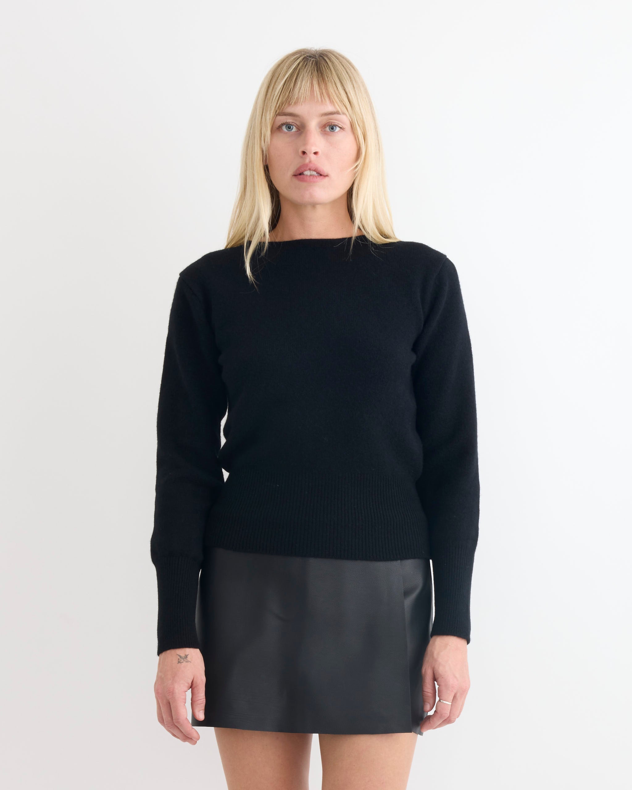 A woman with straight blonde hair wears the Comoli Lambswool Boat Neck Knit, paired with a black leather mini skirt, standing against a white background and looking directly at the camera with a neutral expression.