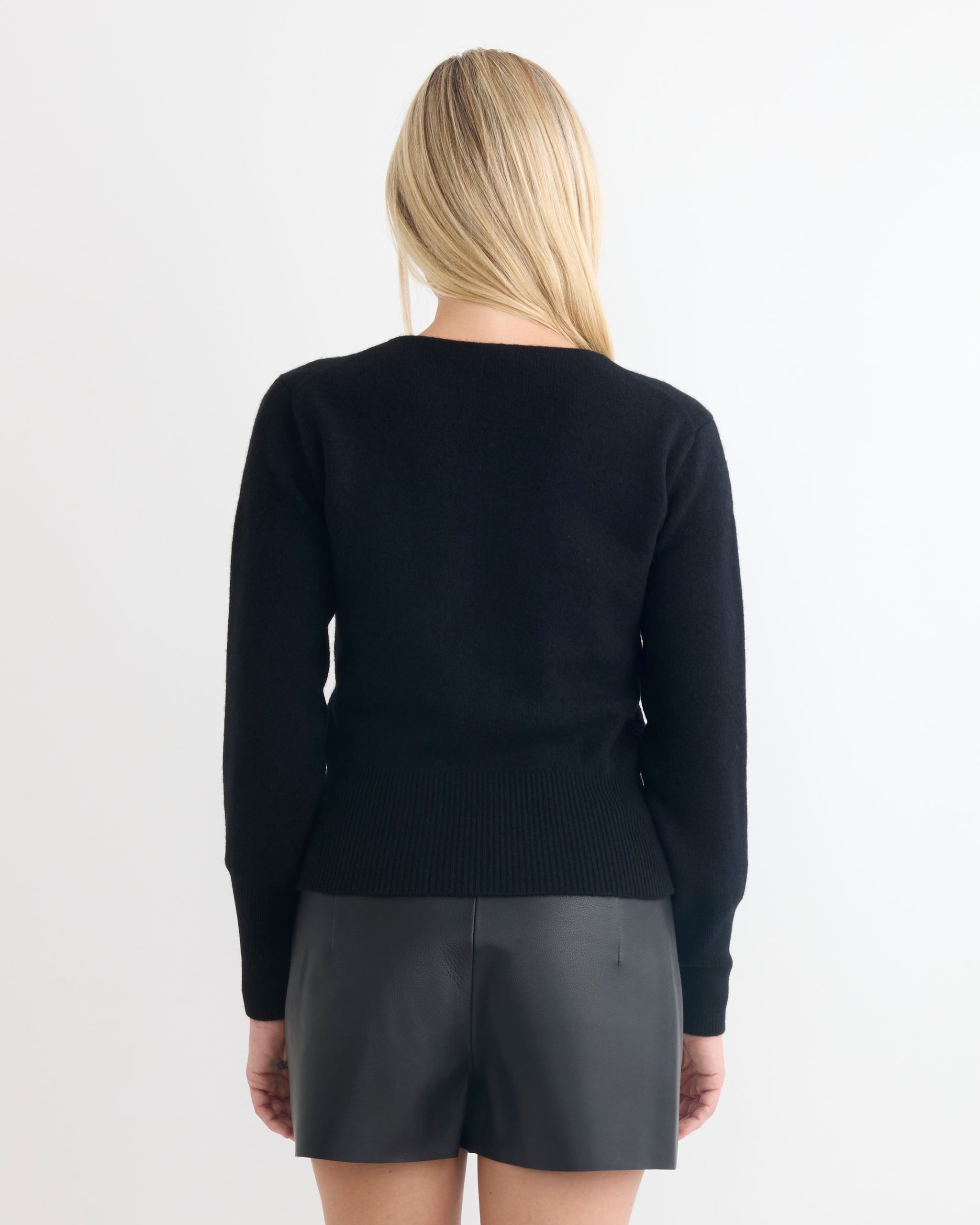 A woman with straight blonde hair stands with her back to the camera, wearing the Comoli Lambswool Boat Neck Knit and black leather shorts against a plain light background.