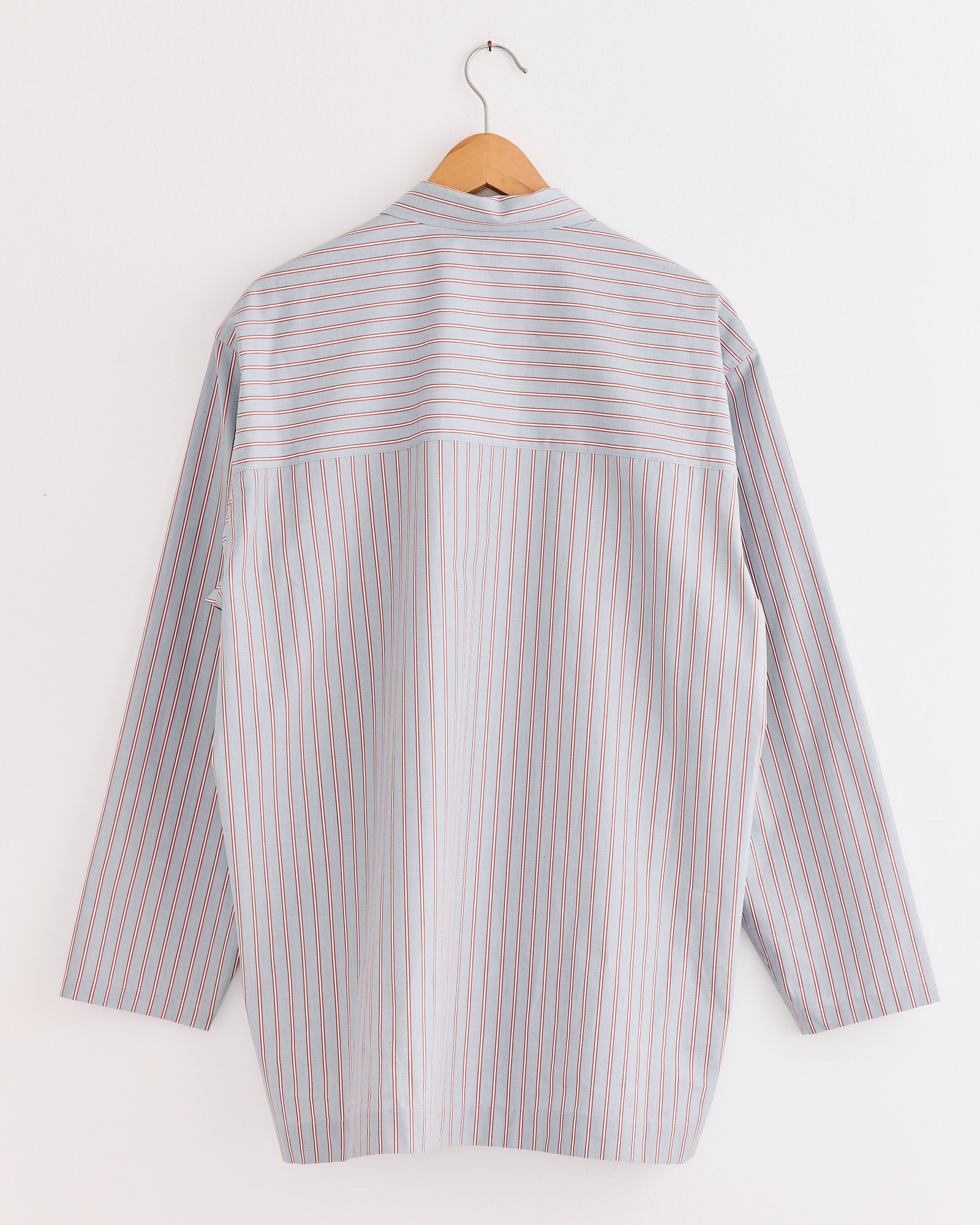 The Cale 80s Stripe Shirt in Blue/Burgundy, a Japanese-made long-sleeve button-up with vertical stripes, is displayed on a wooden hanger against a white wall, shown from the back.