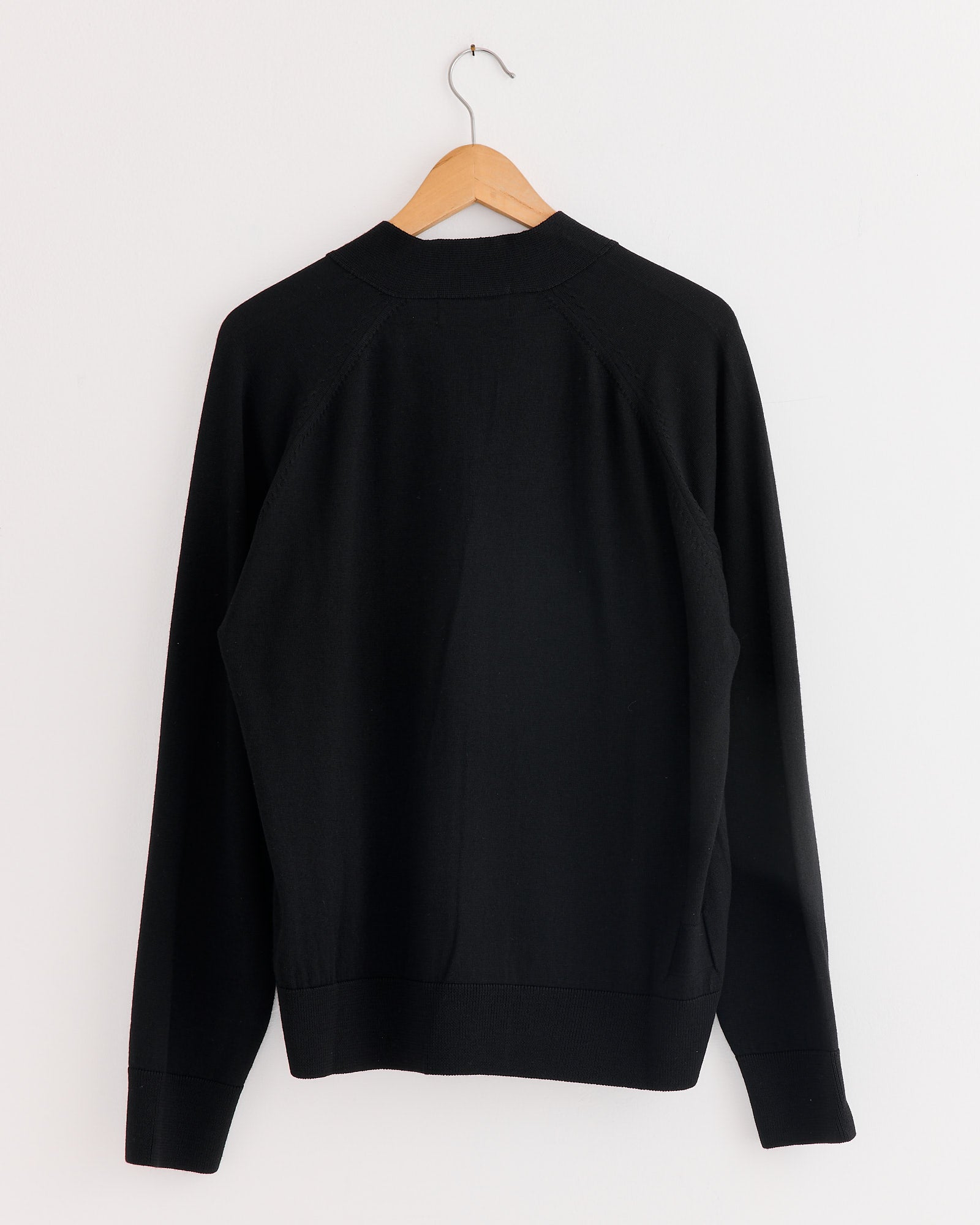 The Loose Cardigan in Black by Universal Works hangs on a wooden hanger against a white wall, displayed from the back.