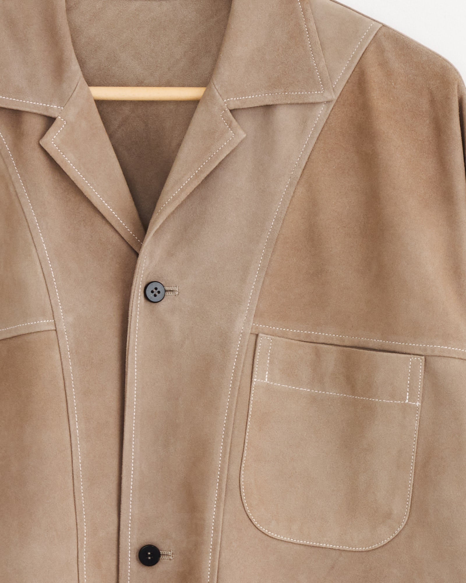 A close-up of the Cale Sheepskin Suede Jacket in Beige on a hanger, showing white stitching, a notched collar, two black buttons, and a large left front pocket. Made in Japan.