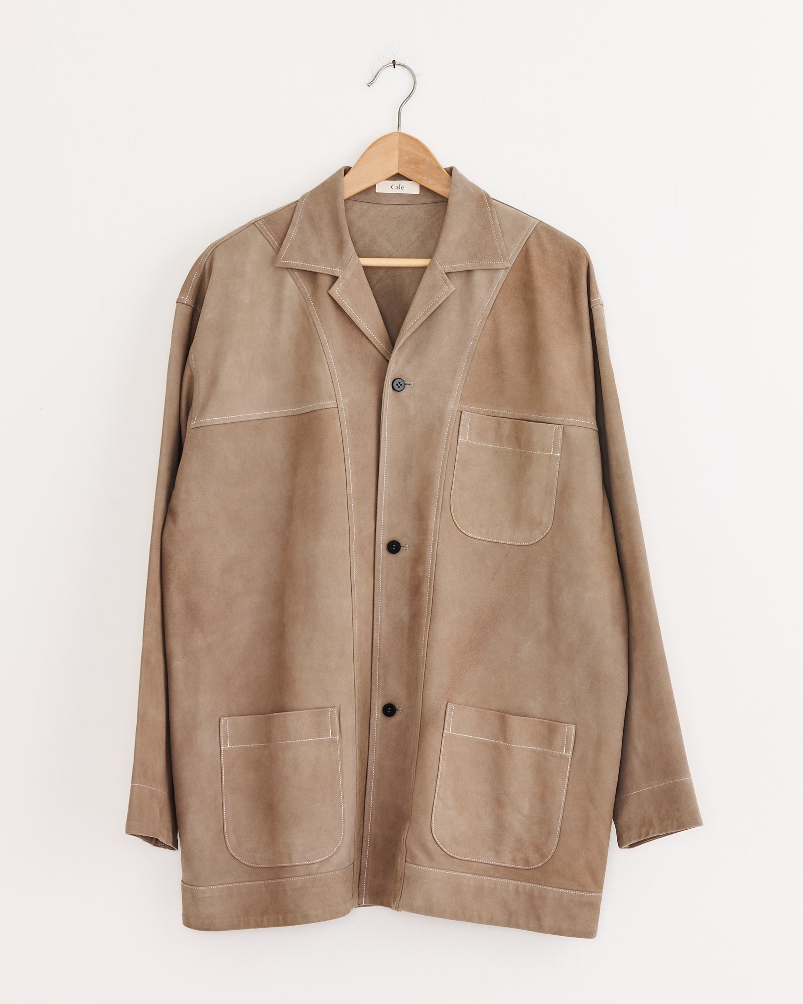 The Cale Sheepskin Suede Jacket in Beige, featuring three large front pockets and white stitching, hangs on a wooden hanger against a plain white wall.