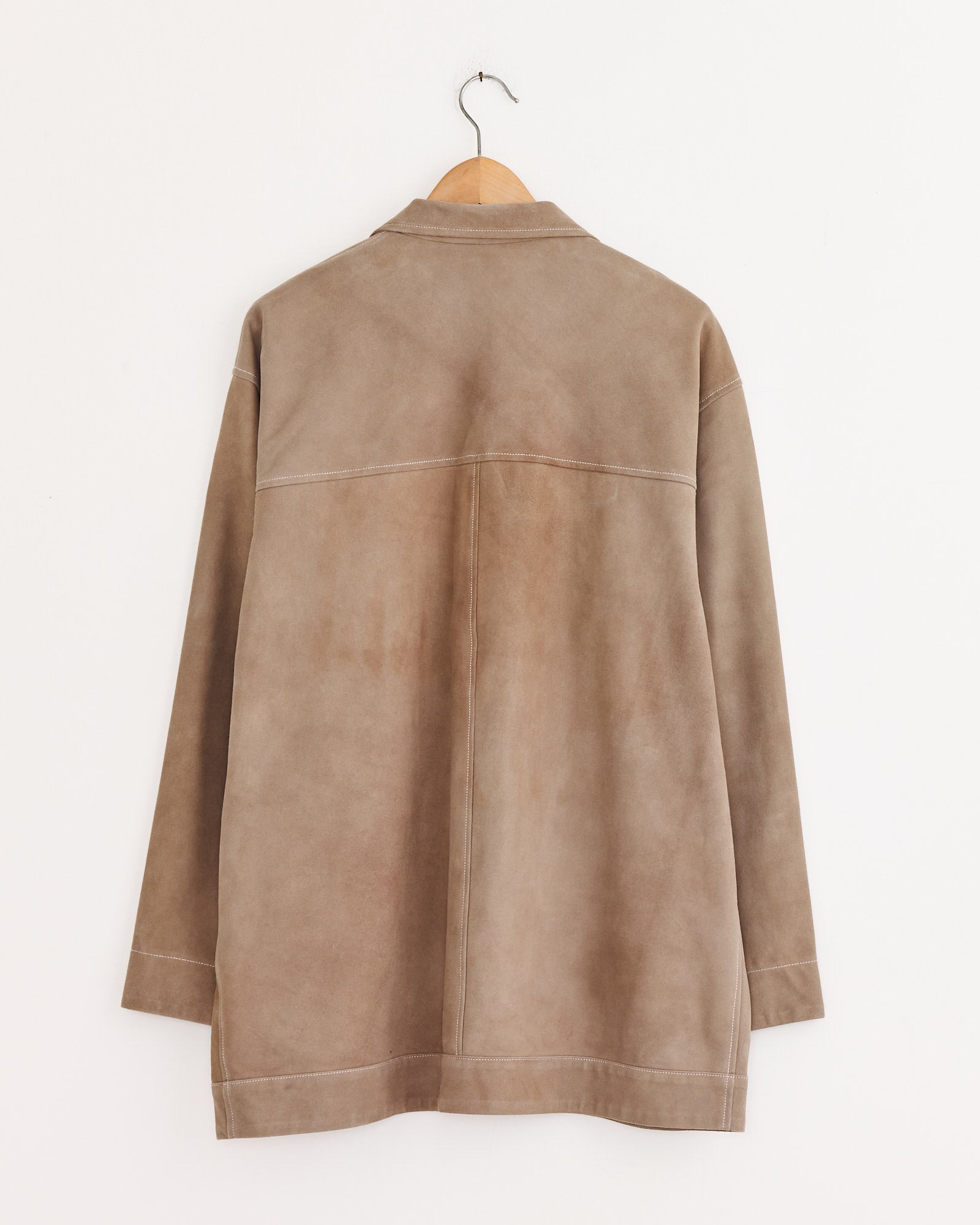 The Cale Sheepskin Suede Jacket in Beige, made in Japan, hangs on a wooden hanger against a plain white wall, displayed from the back to show seam details and long sleeves.