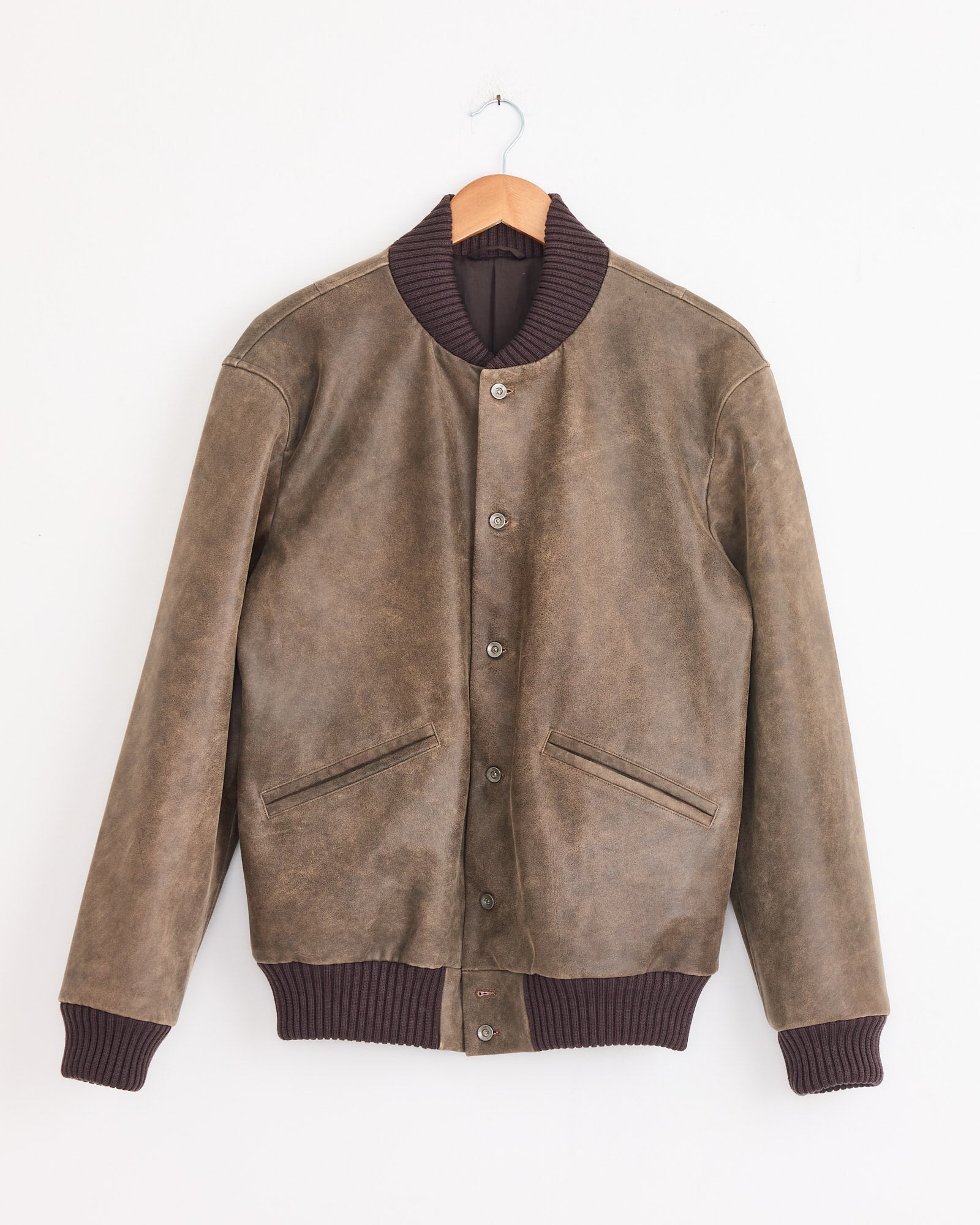 The Cale Antique Calf Jacket in Brown features ribbed dark brown collar, cuffs, and hem, a button-up front, and two slanted pockets. Shown on a wooden hanger against a white wall. Made in Japan.