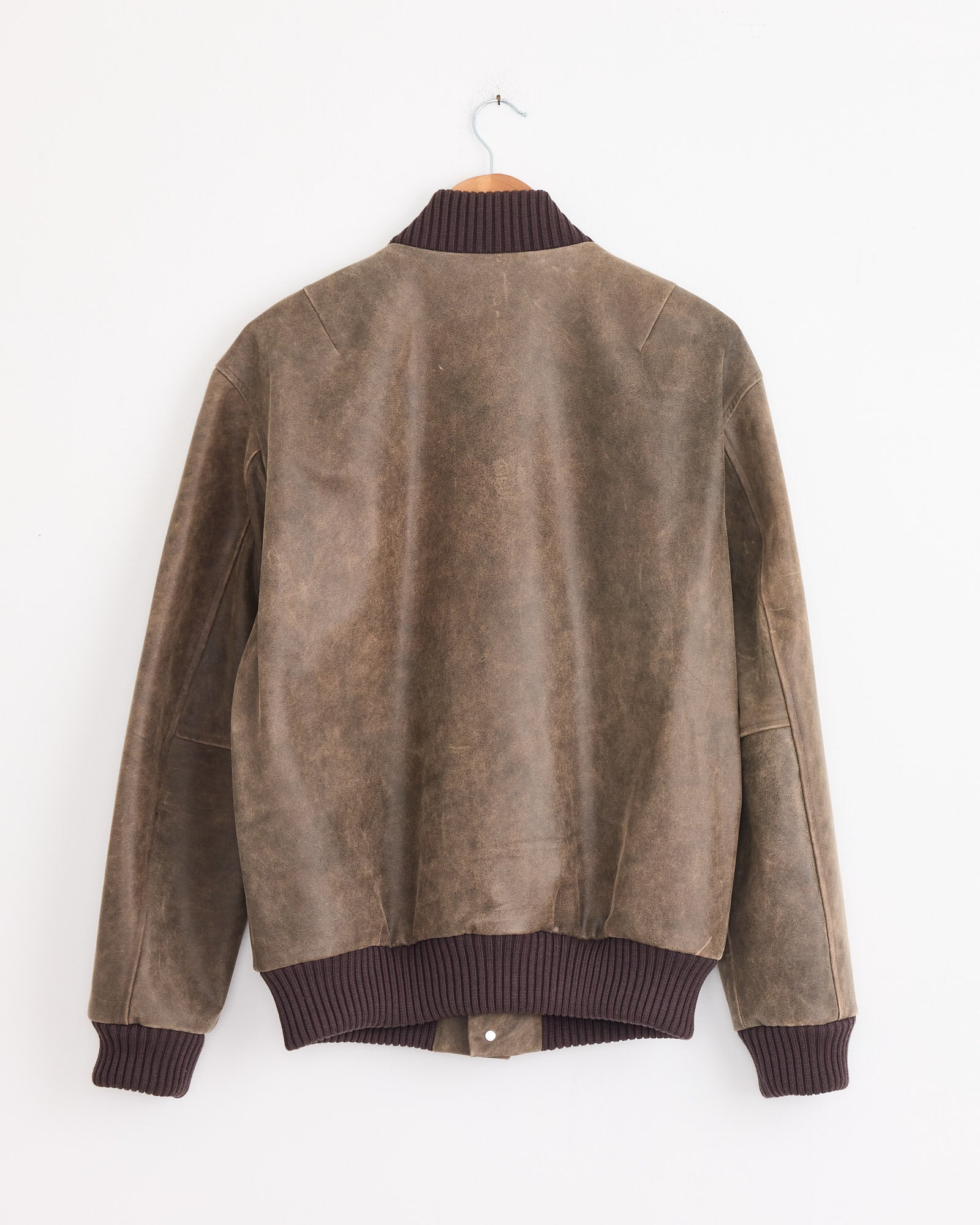 The Antique Calf Jacket in Brown by Cale, featuring dark ribbed knit cuffs, waistband, and collar, is displayed from the back on a wooden hanger against a white wall.
