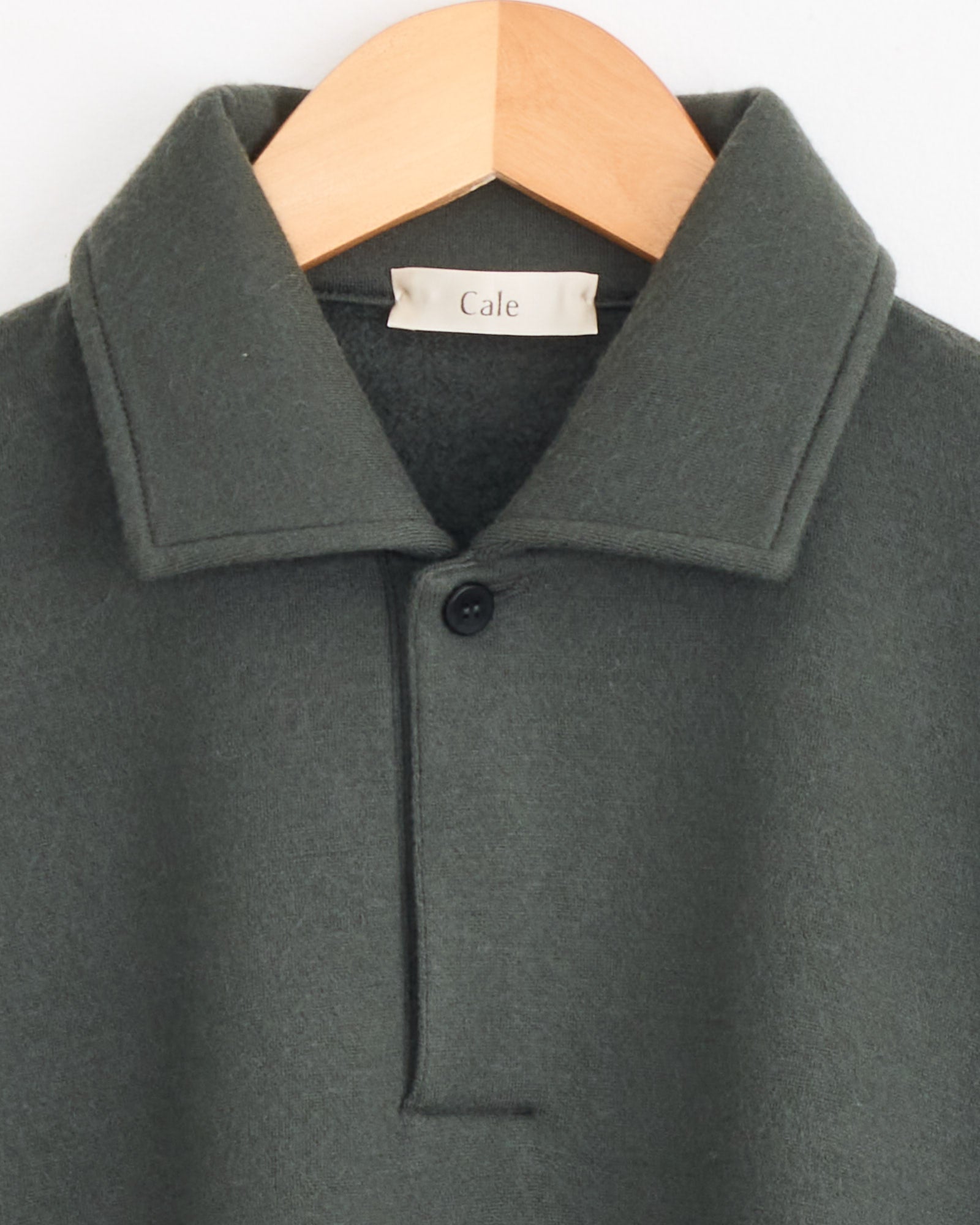 A close-up of the Cale 120 Air Wool Fleece Polo in khaki, featuring a single black button and “Cale” label, displayed on a wooden hanger.