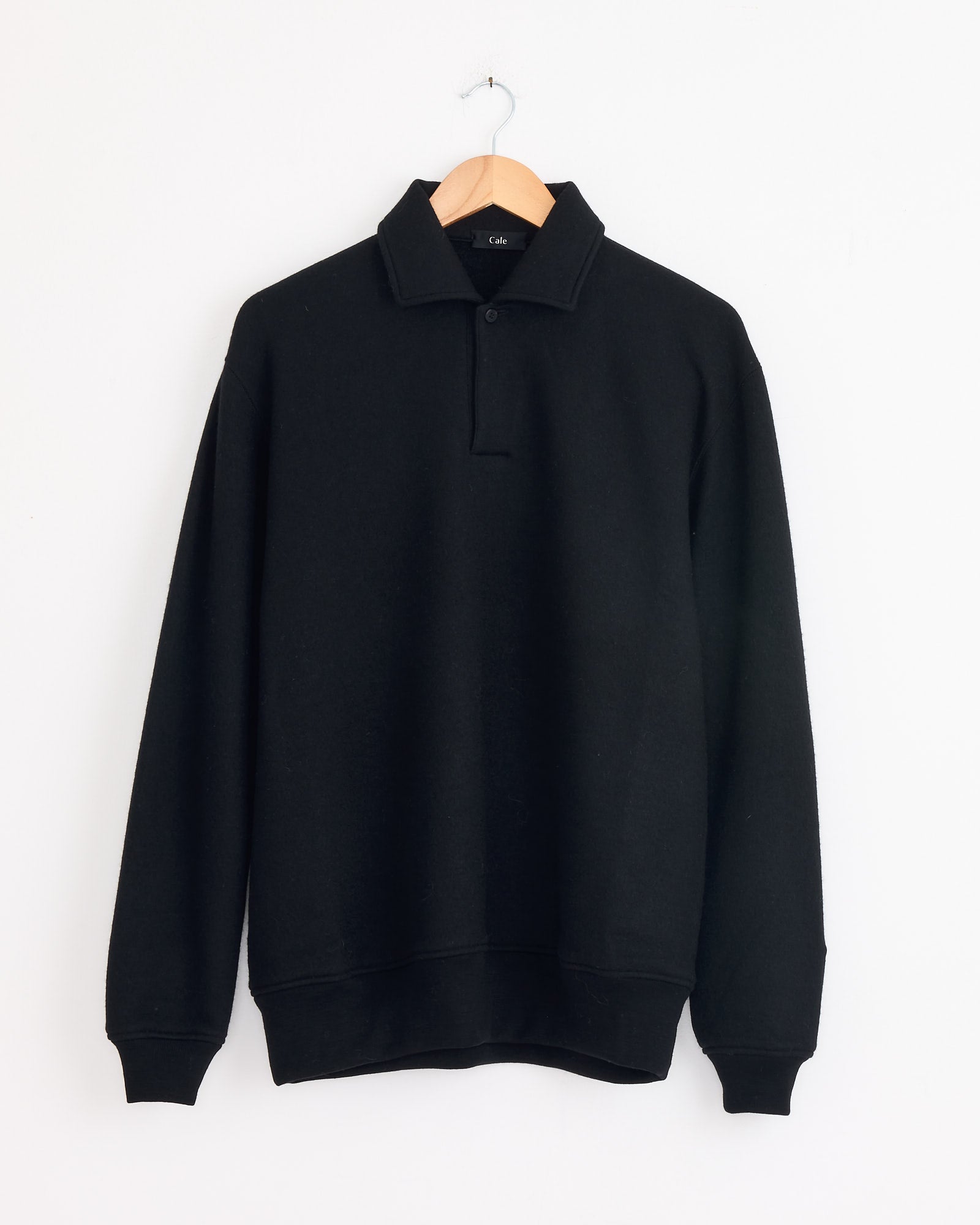 The Cale 120 Air Wool Fleece Polo in Black, featuring a collar and two buttons, made in Japan, hangs on a wooden hanger against a plain white wall.