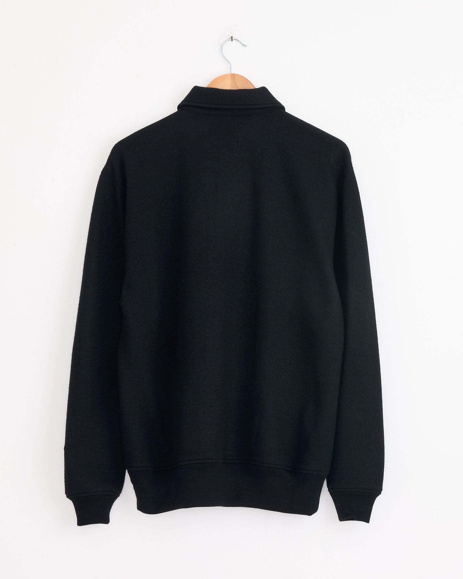The Cale 120 Air Wool Fleece Polo in Black, crafted from Super 120's wool and made in Japan, features long sleeves and is shown from the back hanging on a wooden hanger against a white wall.