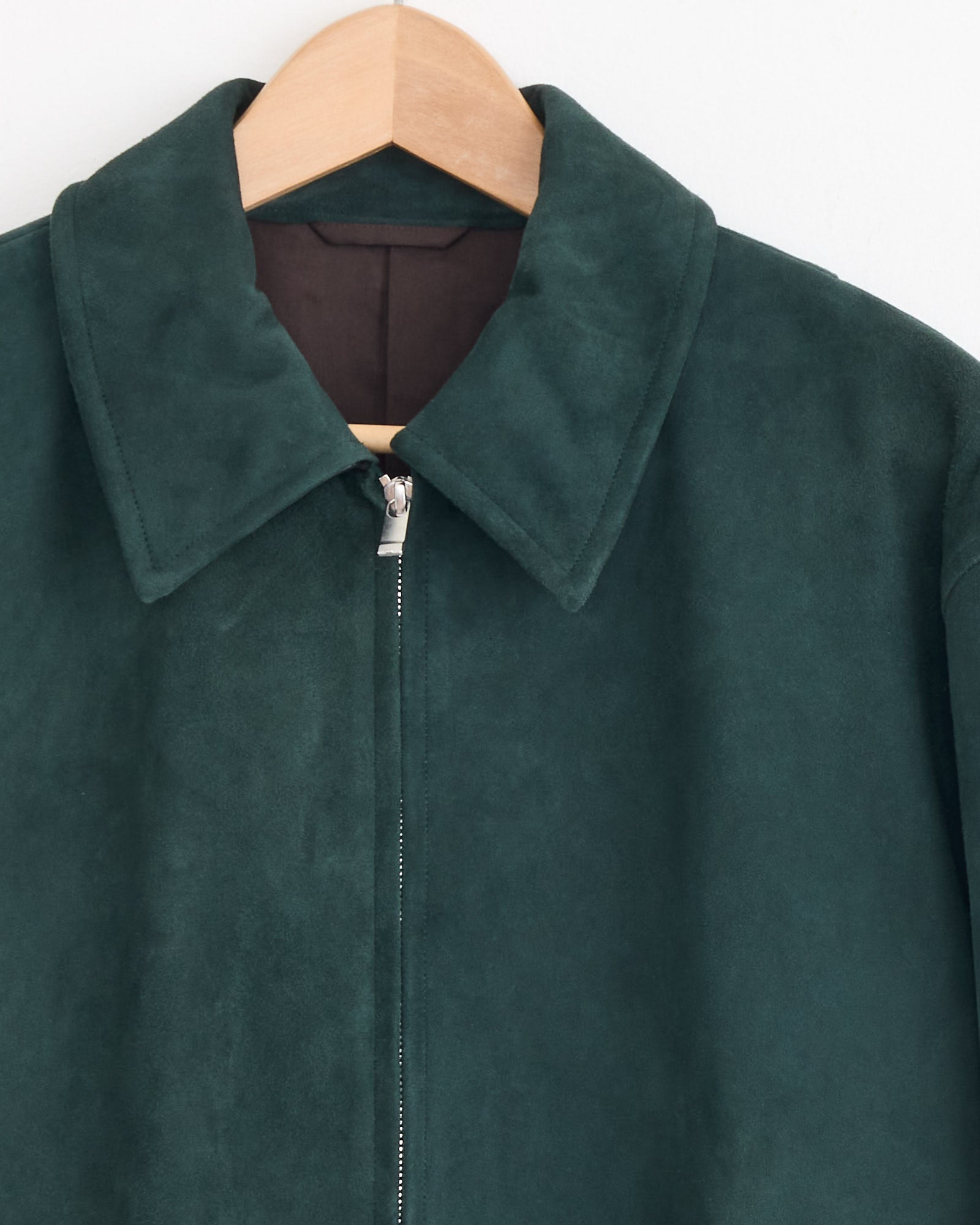 The Cale Sheep Suede Heavy Jacket in Green, made in Japan, features a wide collar, smooth surface, and silver zipper. Displayed on a wooden hanger against a white wall.