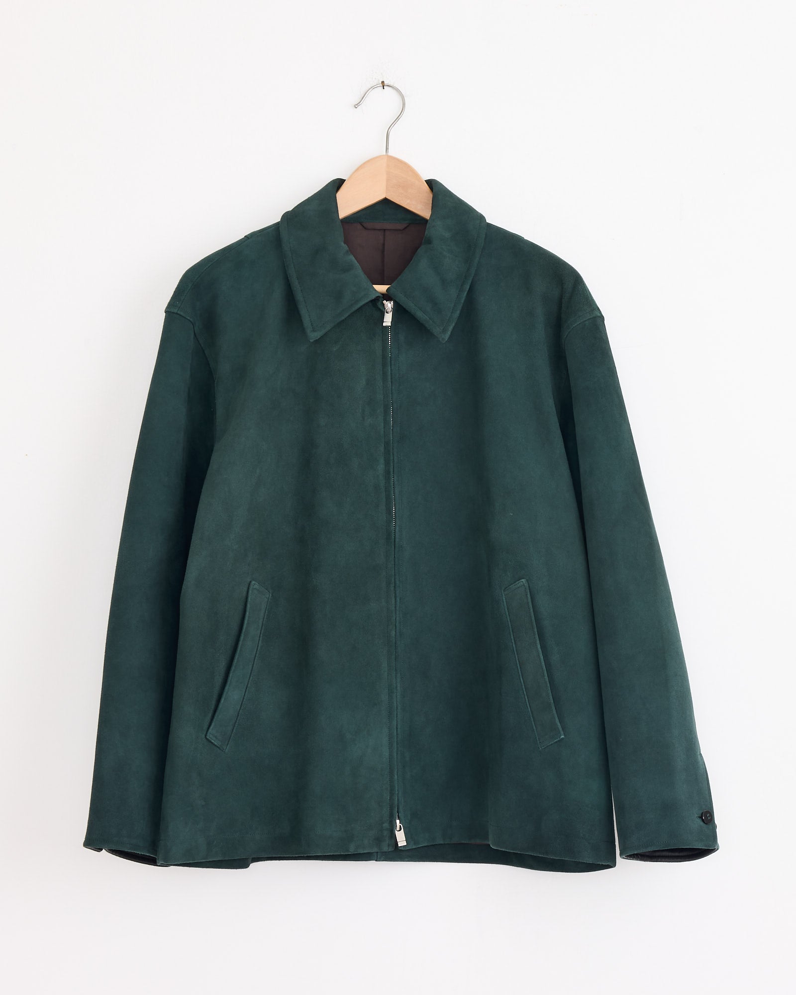 The Cale Sheep Suede Heavy Jacket in Green features a collar, front zipper, and two slanted pockets. Expertly made in Japan, it's shown on a wooden hanger against a plain white wall for a minimalist look.