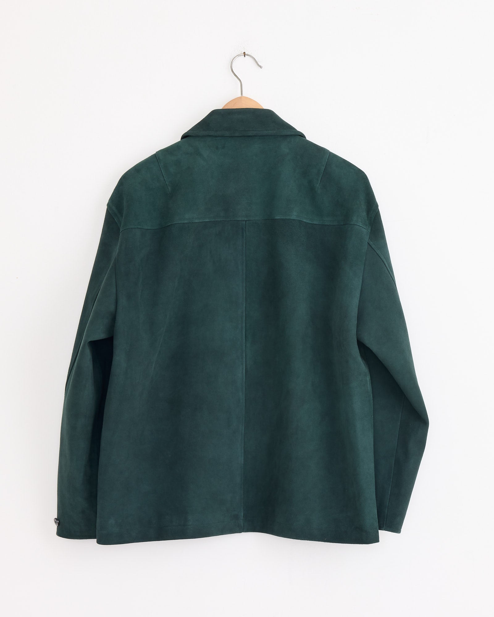 The Cale Sheep Suede Heavy Jacket in Green hangs on a wooden hanger against a white wall, shown from the back with long sleeves and a simple collar. Made in Japan, it showcases timeless style and superior craftsmanship.