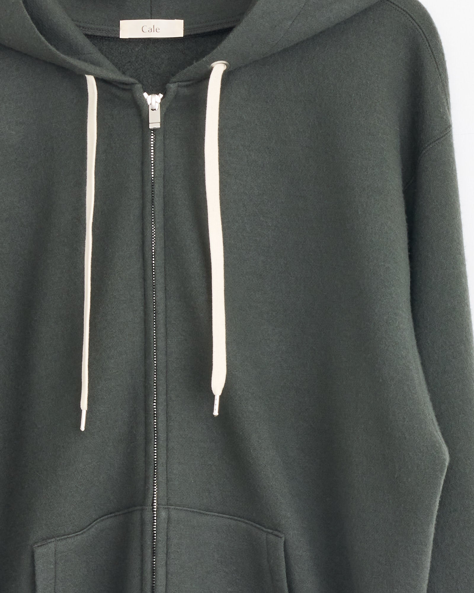 The Cale 120 Air Wool Fleece Hoodie in Khaki is a dark green zip-up hoodie made in Japan from soft wool fleece. It features white drawstrings, a front zipper, a relaxed fit, and a visible Cale label inside the collar.
