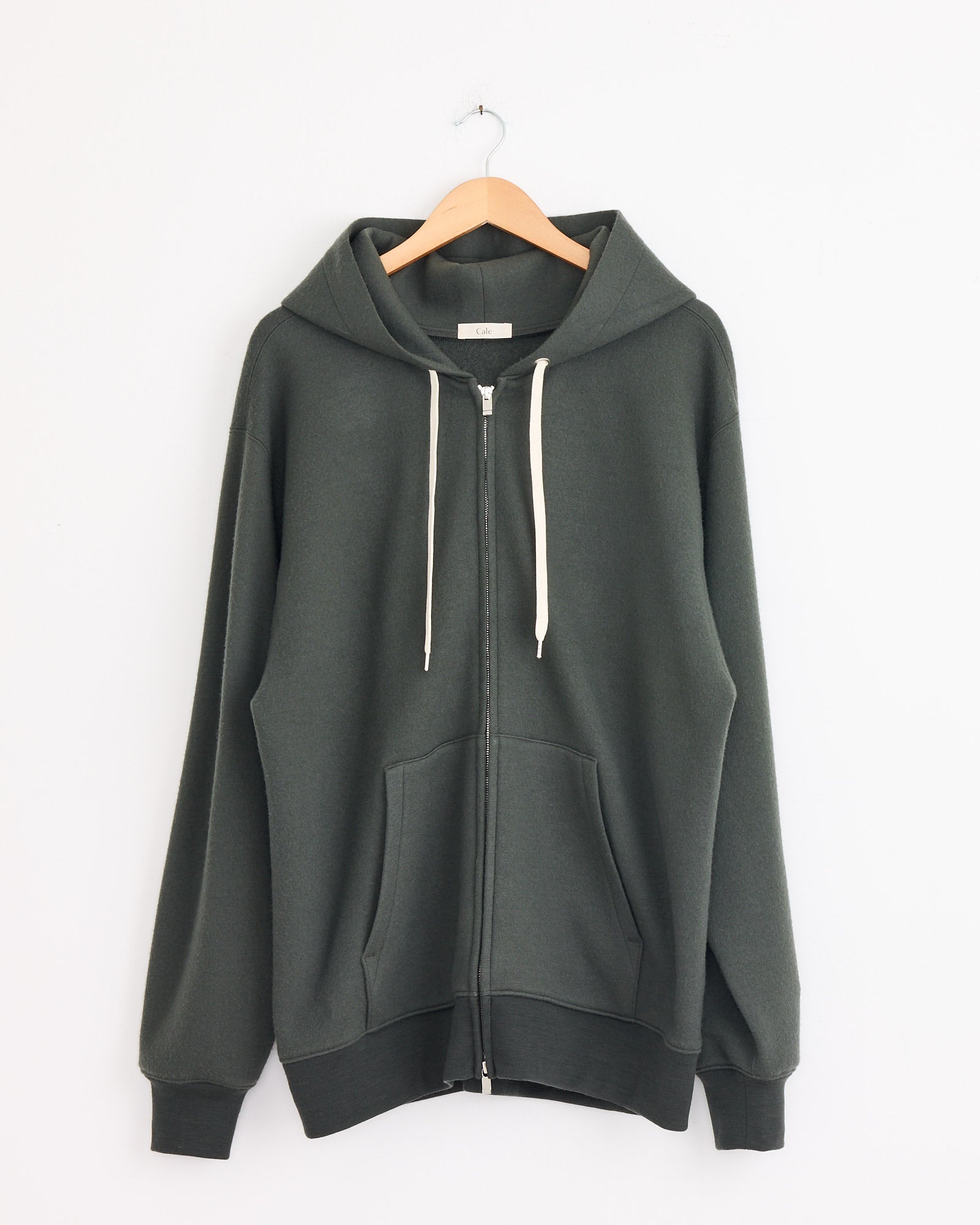 The Cale 120 Air Wool Fleece Hoodie in Khaki hangs on a wooden hanger against a white wall, featuring front pockets, ribbed cuffs and hem, a zip-up design with white drawstrings. Made in Japan.