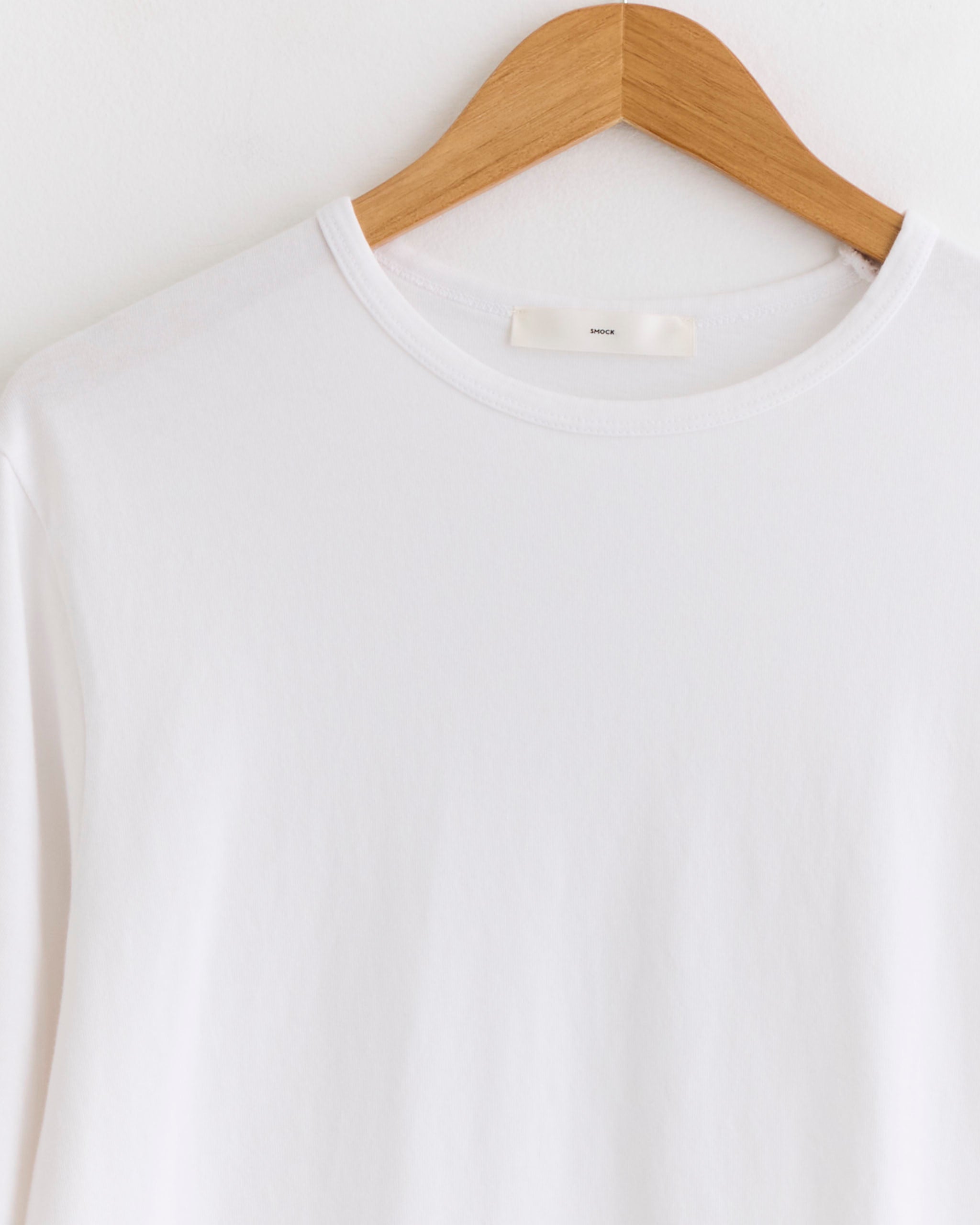 The SMOCK Under Tee in White, made from combed cotton, hangs on a wooden hanger against a white background—ideal as an underlayer.