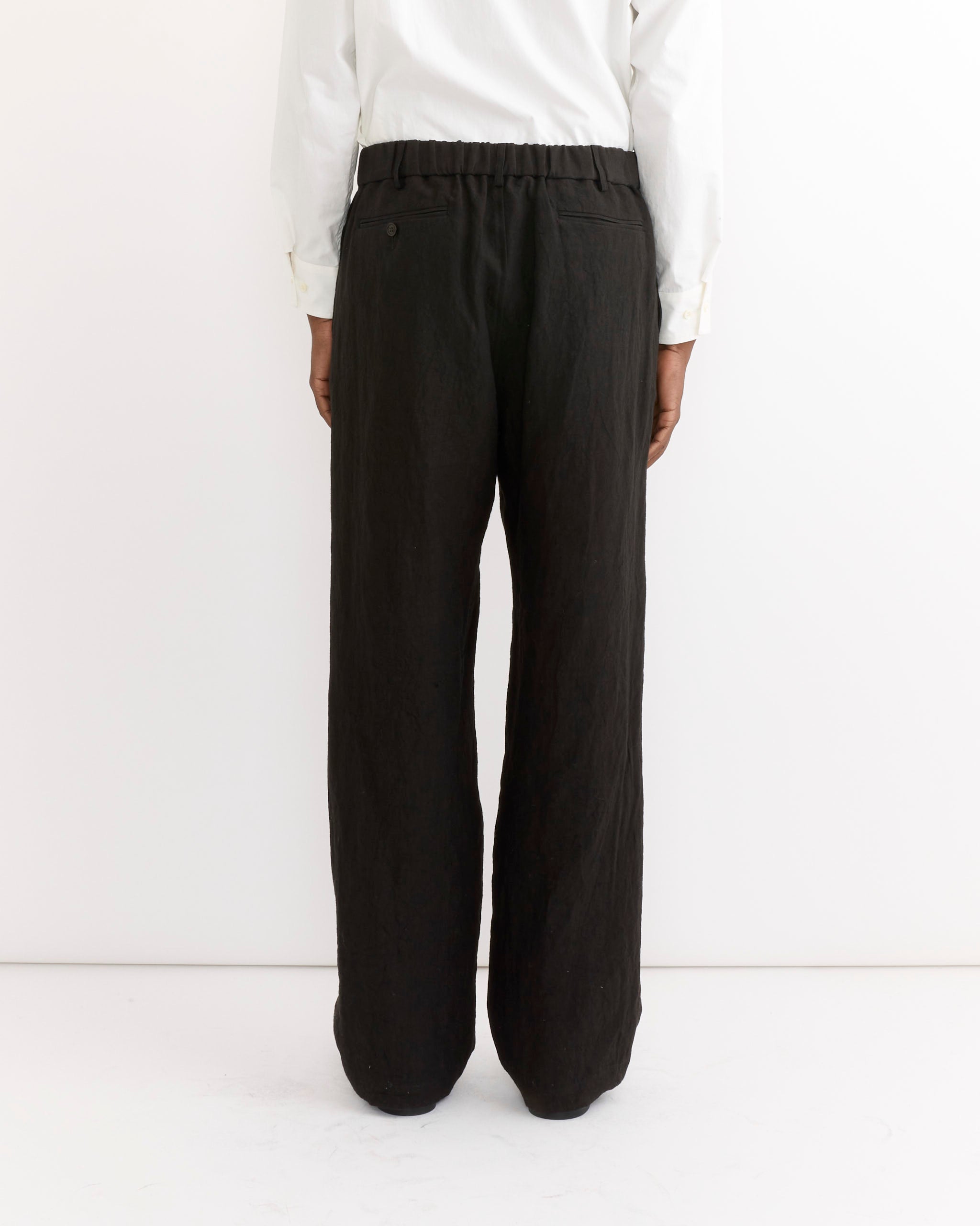 A person stands with their back to the camera, wearing a white long-sleeve shirt and SMOCK's West Coast Pant in Sun Dried Linen Black, posed against a plain white background.