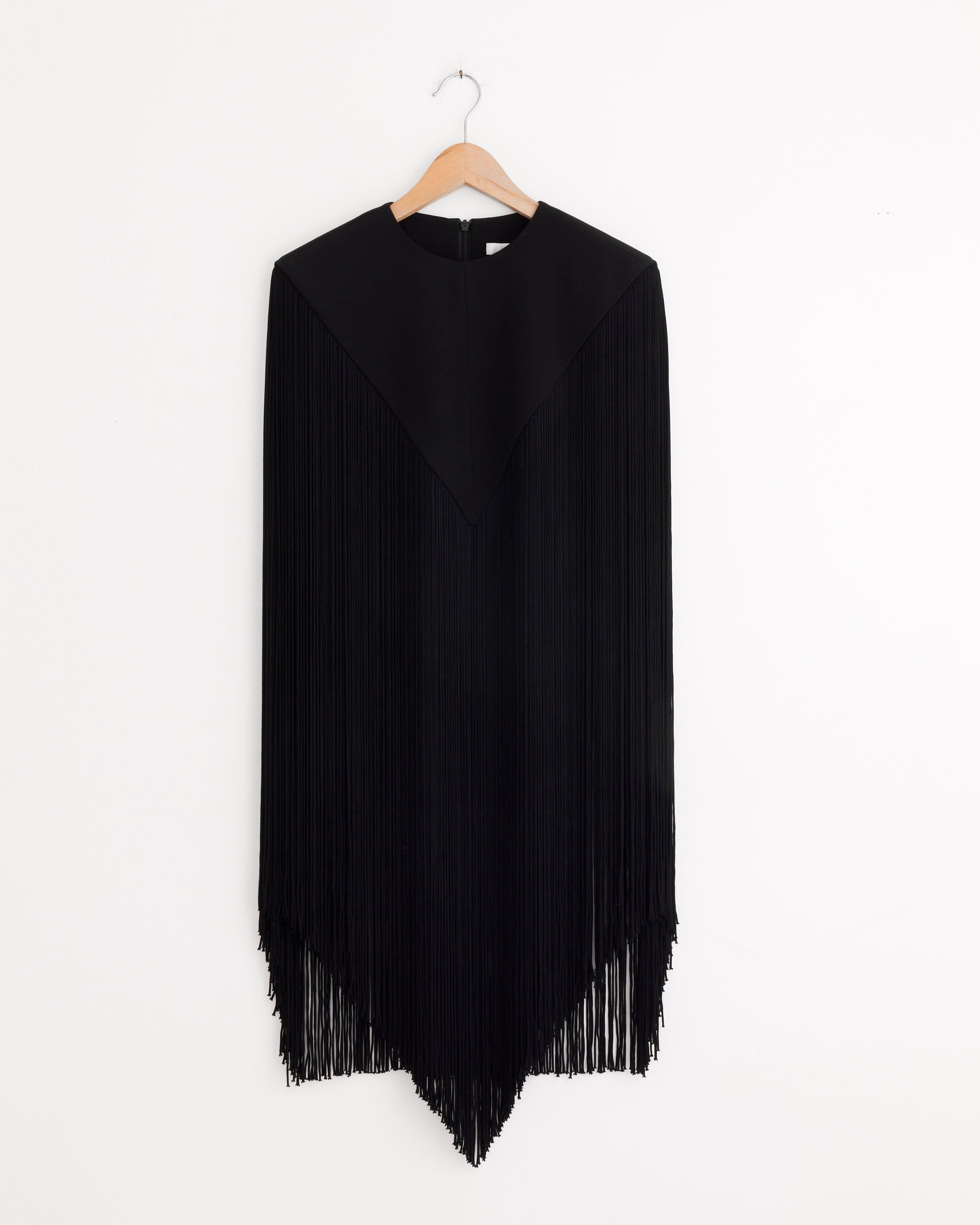 The Tibi 4-Ply Silk Fringe Cape in Black features a pointed hem and long fringe, offering the perfect lightweight silk layer for evening elegance. Shown on a wooden hanger against a plain white background.